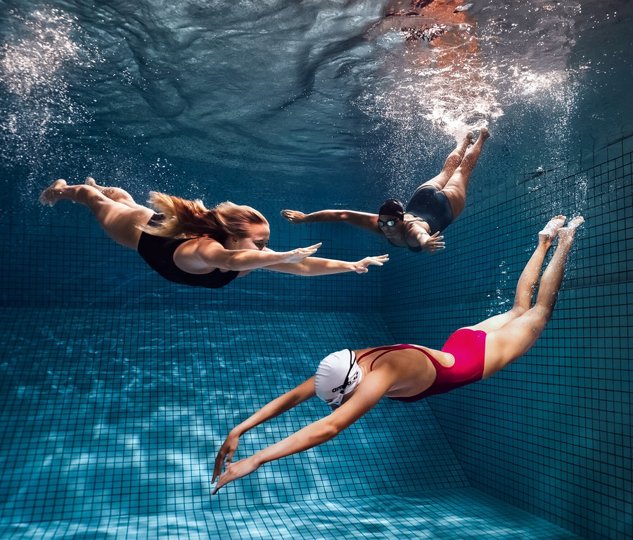 Drei Frauen schwimmen unter Wasser in einem Schwimmbad. Eine trägt einen arena Shapewear Damen Badeanzug Lara C-Cup.