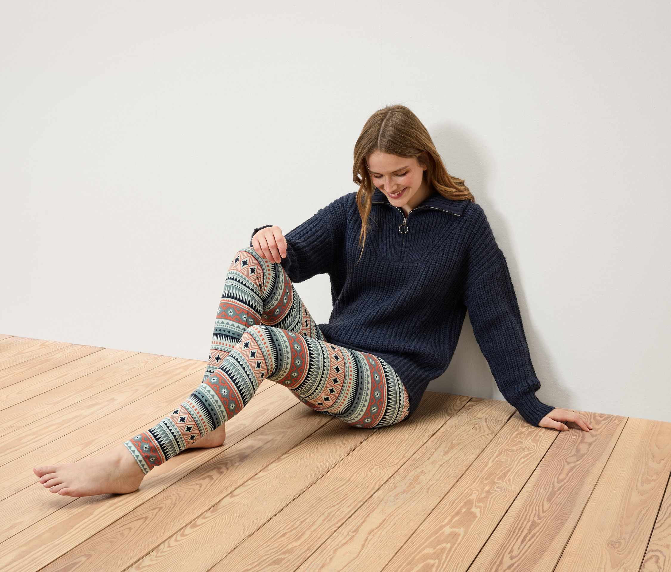 Frau sitzt auf einem Holzboden und trägt einen dunkelblauen Pullover mit Reißverschluss und gemusterte Leggings.
