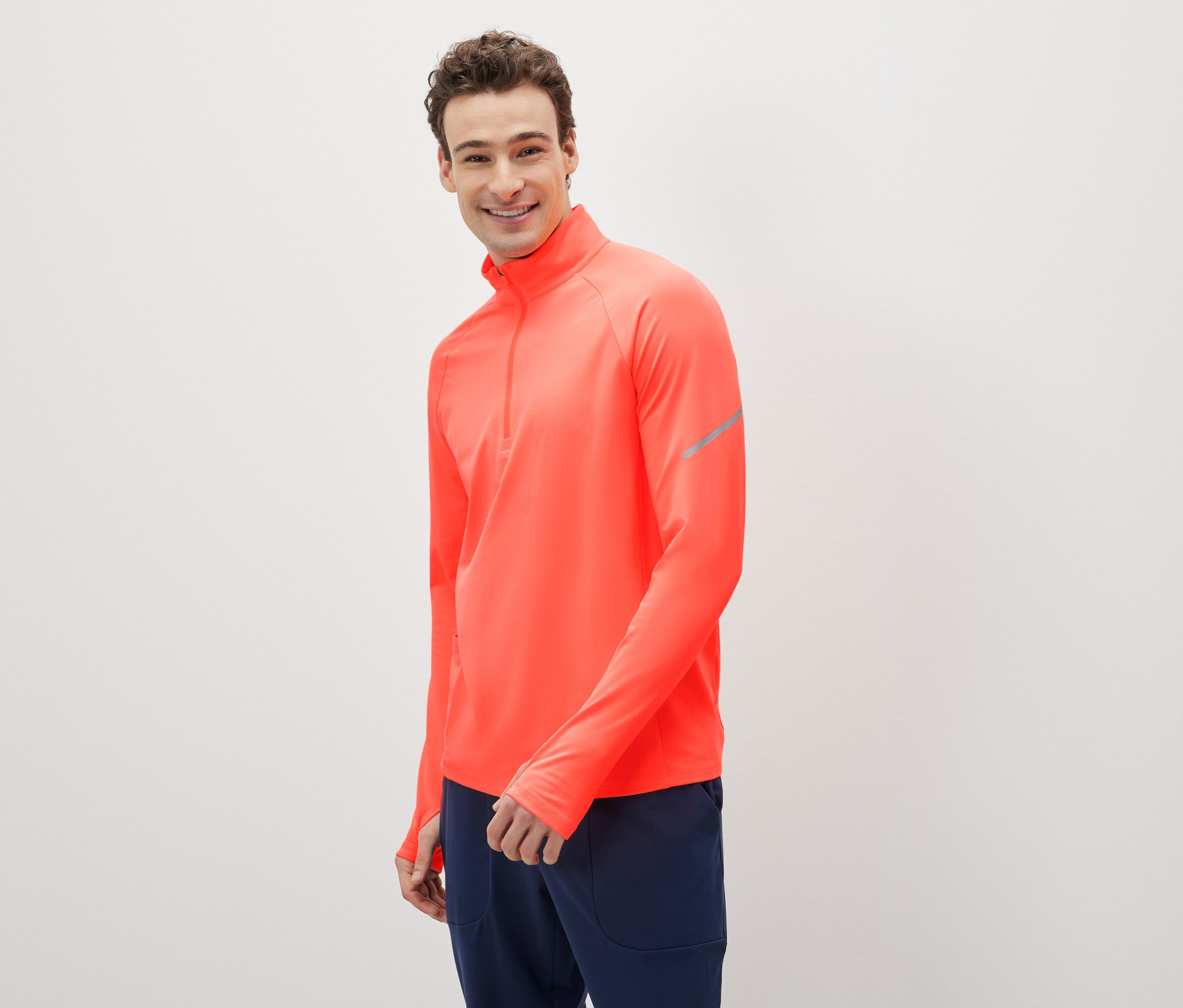 Ein Mann lächelt und posiert in einem korallroten, langärmligen Sportoberteil und dunkelblauen Trainingshosen.