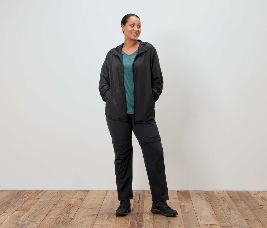 Eine Frau steht mit den Händen in den Taschen mit schwarzer Regenjacke, grünem Oberteil und schwarzer Hose vor einer weißen Wand.