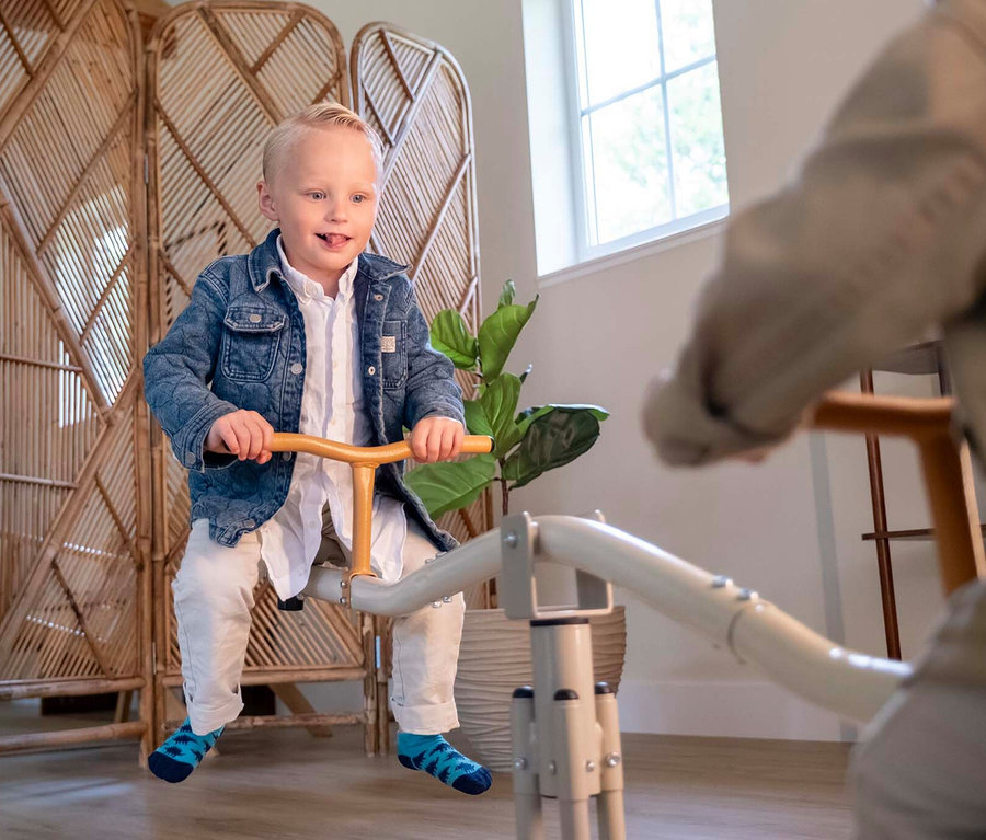Ein kleiner Junge mit blauen Socken sitzt auf einer grauen Wippe mit Holzgriffen. Im Hintergrund sind ein Raumteiler und eine Zimmerpflanze zu sehen.