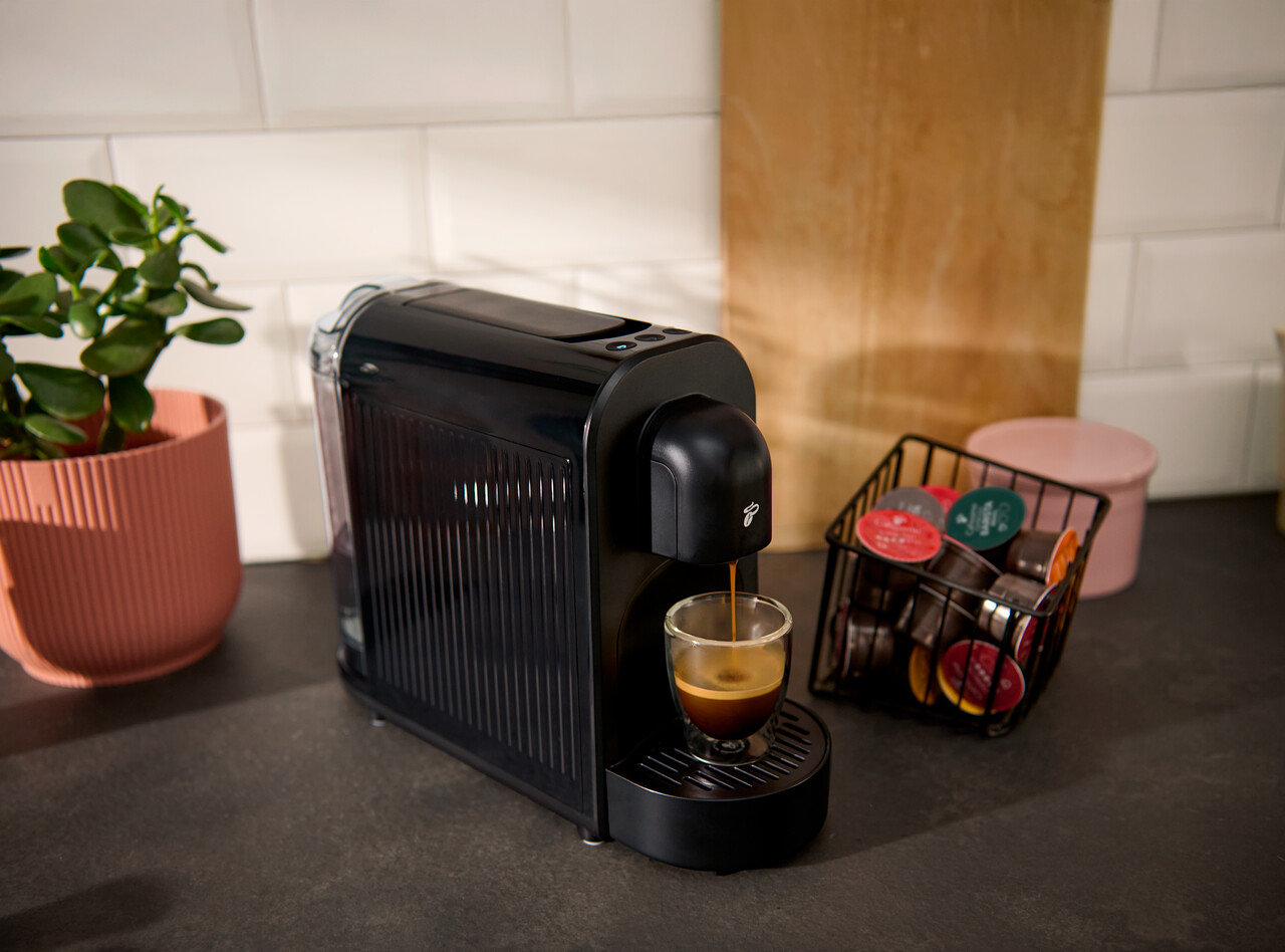 Eine schwarze Kaffeemaschine gießt Kaffee in ein Glas. Daneben stehen Kaffeekapseln in einem Drahtkorb und eine Pflanze in einem Topf.