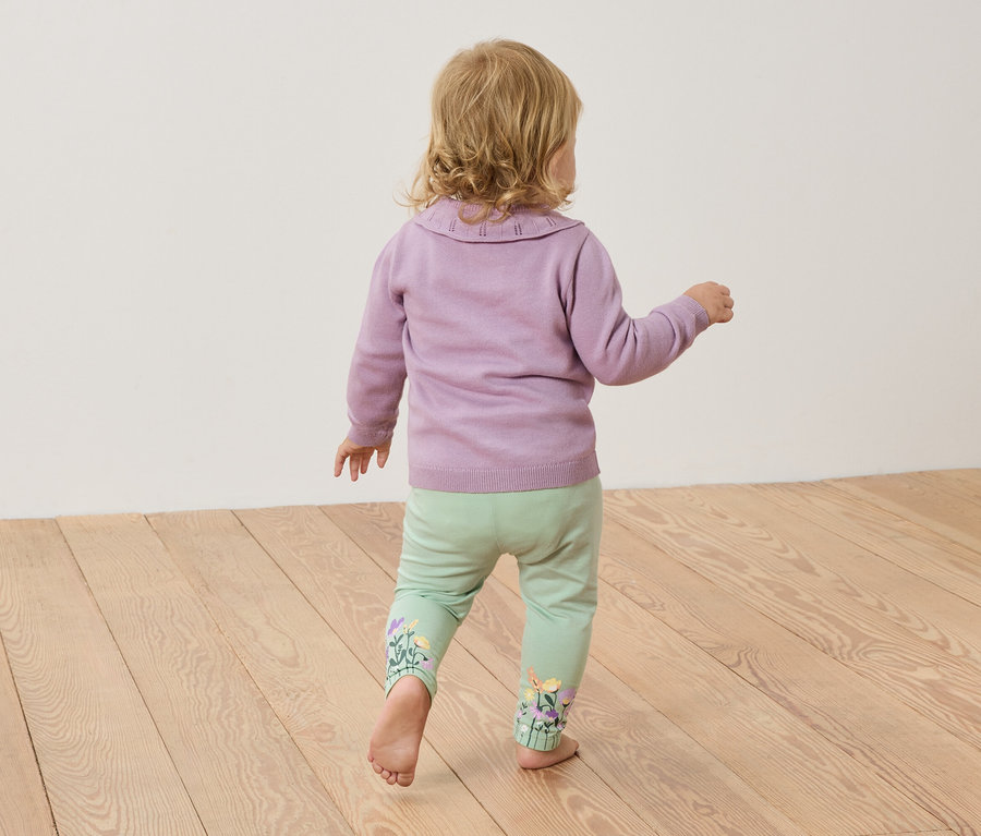 Ein Kleinkind mit blonden Haaren steht barfuß auf einem Holzboden, trägt einen lila Pullover und hellgrüne Hosen mit Blumenstickereien.