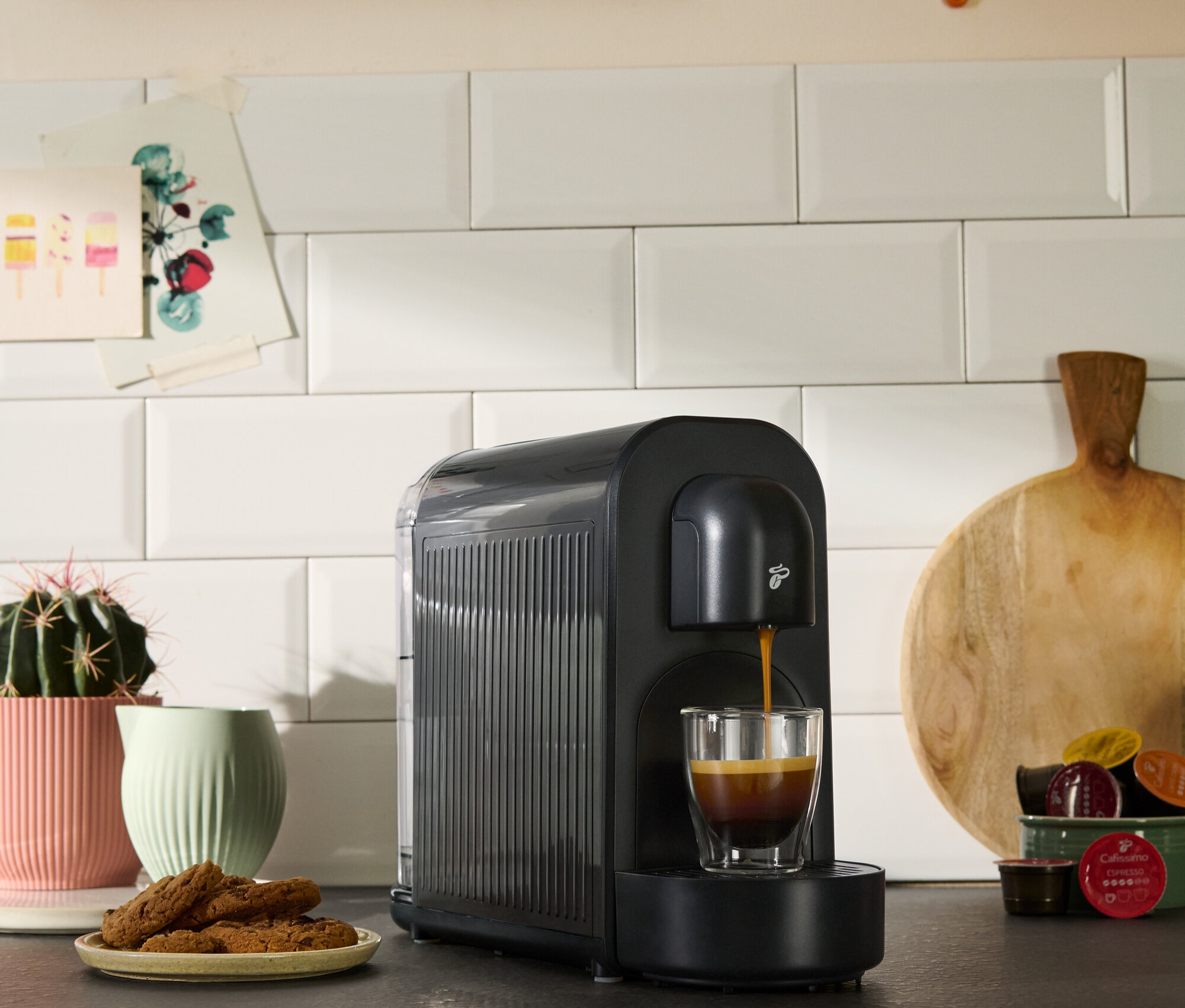 Eine schwarze Kaffeemaschine füllt eine Tasse mit Kaffee. Kekse, eine Sukkulente und Küchendekoration sind ebenfalls zu sehen.