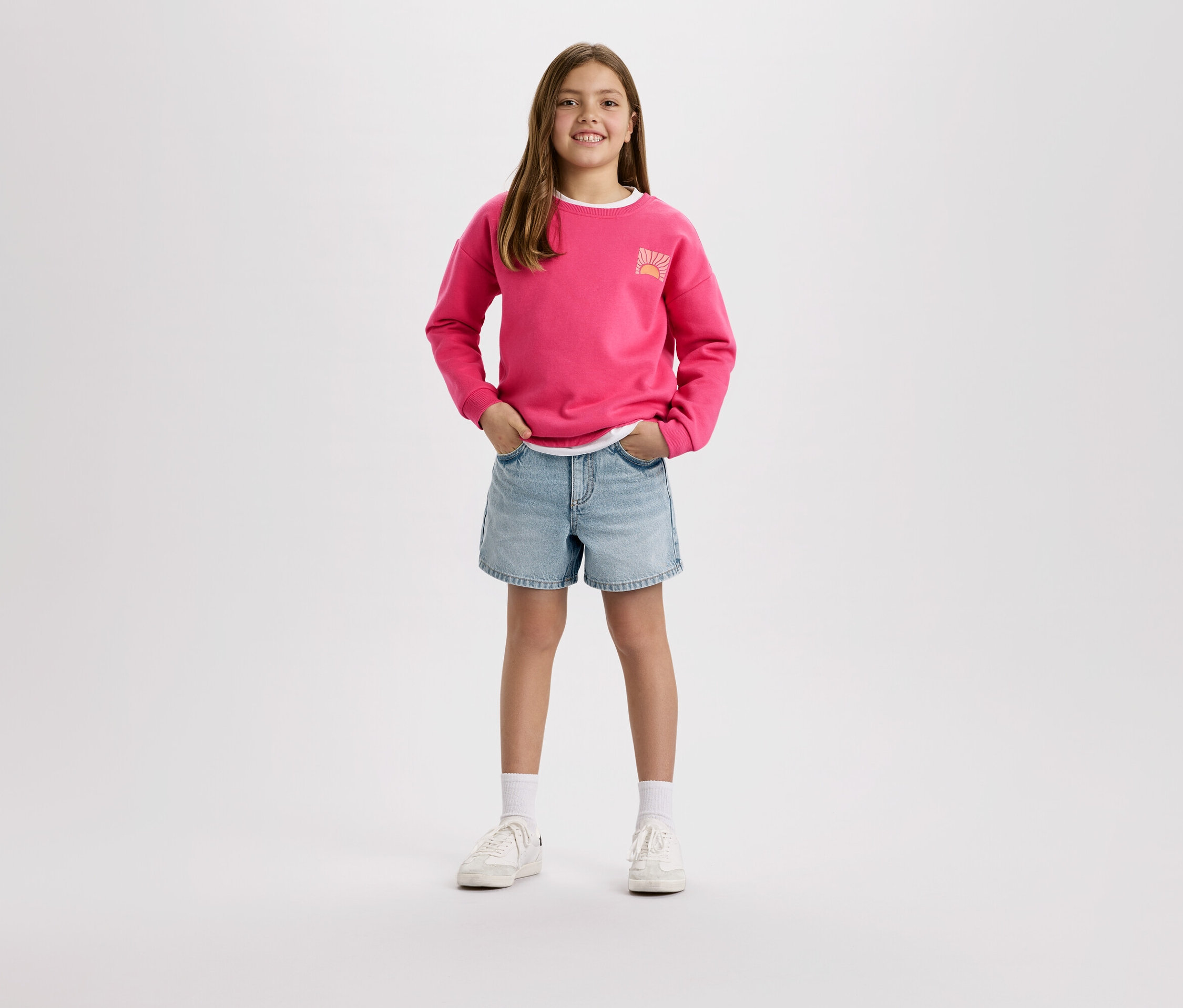 Mädchen mit pinkem Pullover und Jeansshorts steht vor einem weißen Hintergrund.