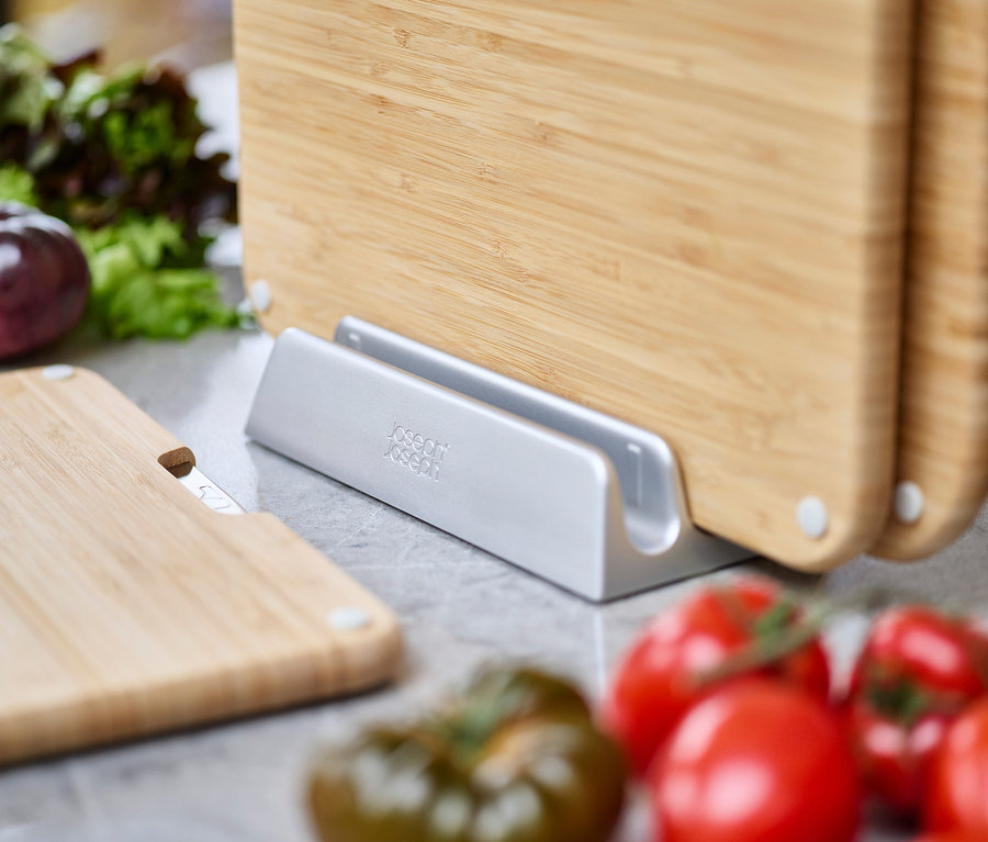 Mehrere Bambus-Schneidebretter stehen in einem silbernen Halter, umgeben von Tomaten und Salat.