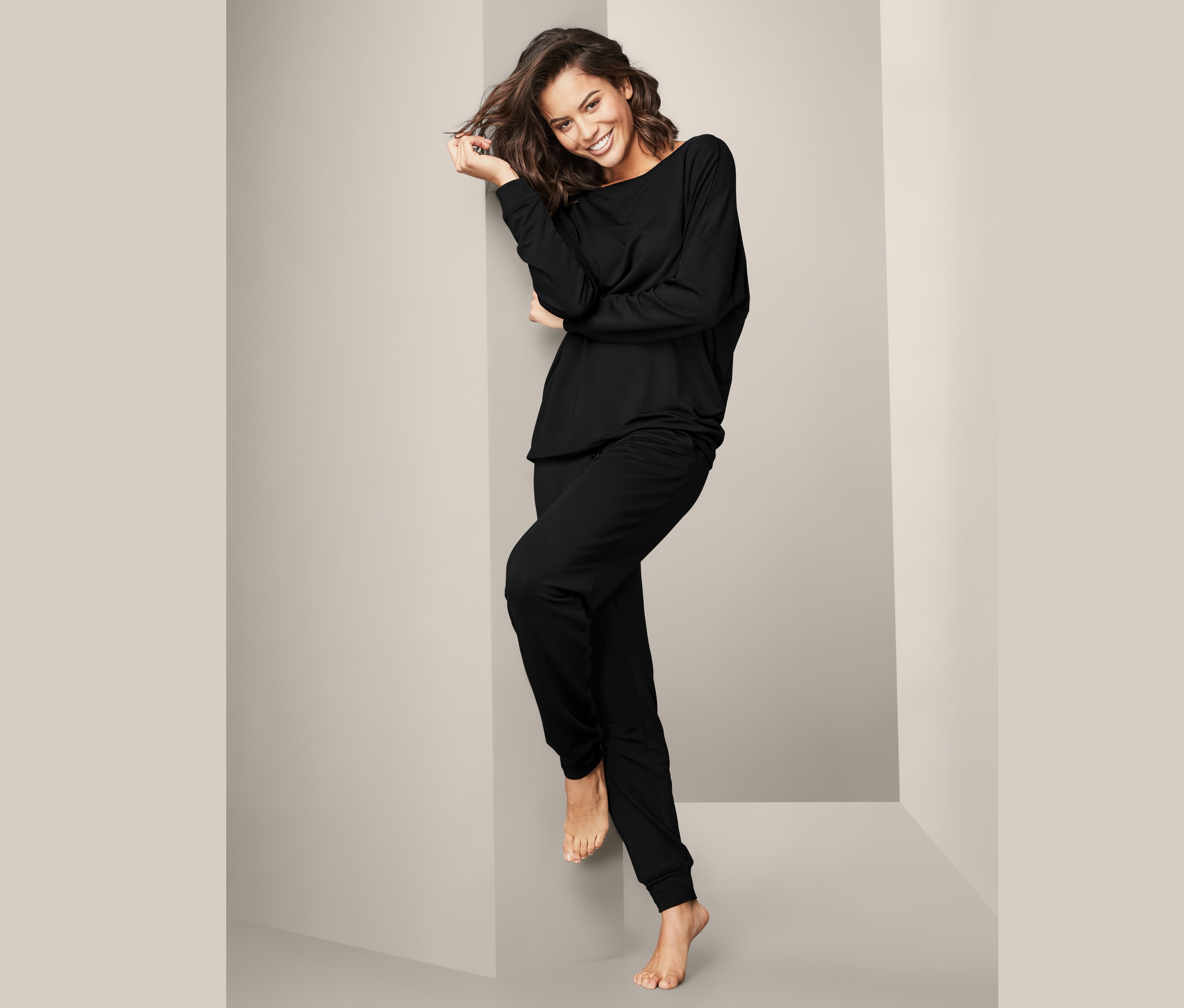 Eine Frau lächelt und lehnt an einer Wand, sie trägt einen schwarzen Hausanzug.