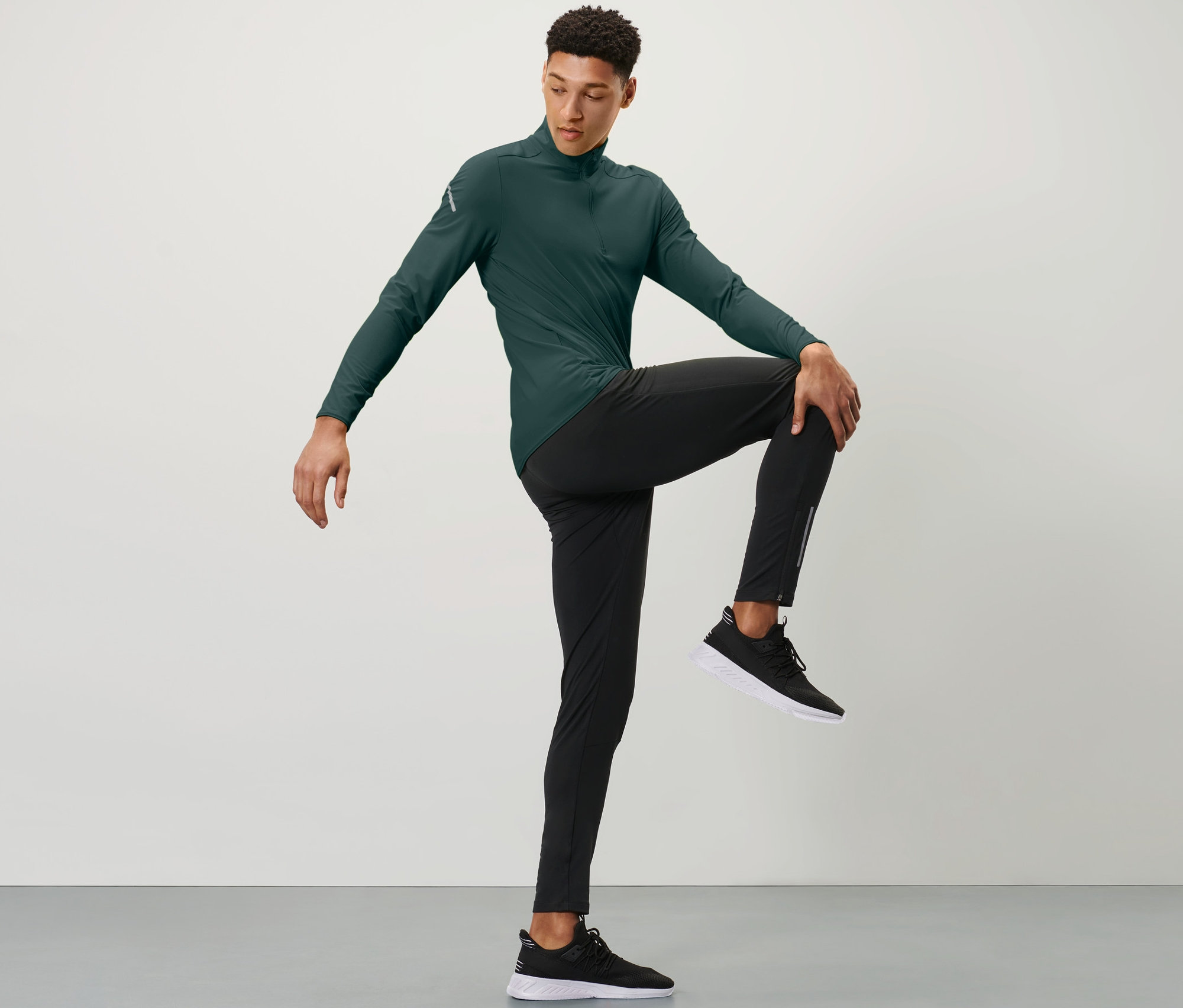 Mann in grünem Sportoberteil und schwarzen Windprotection-Lauftight hält ein Knie mit einer Hand vor der Brust.