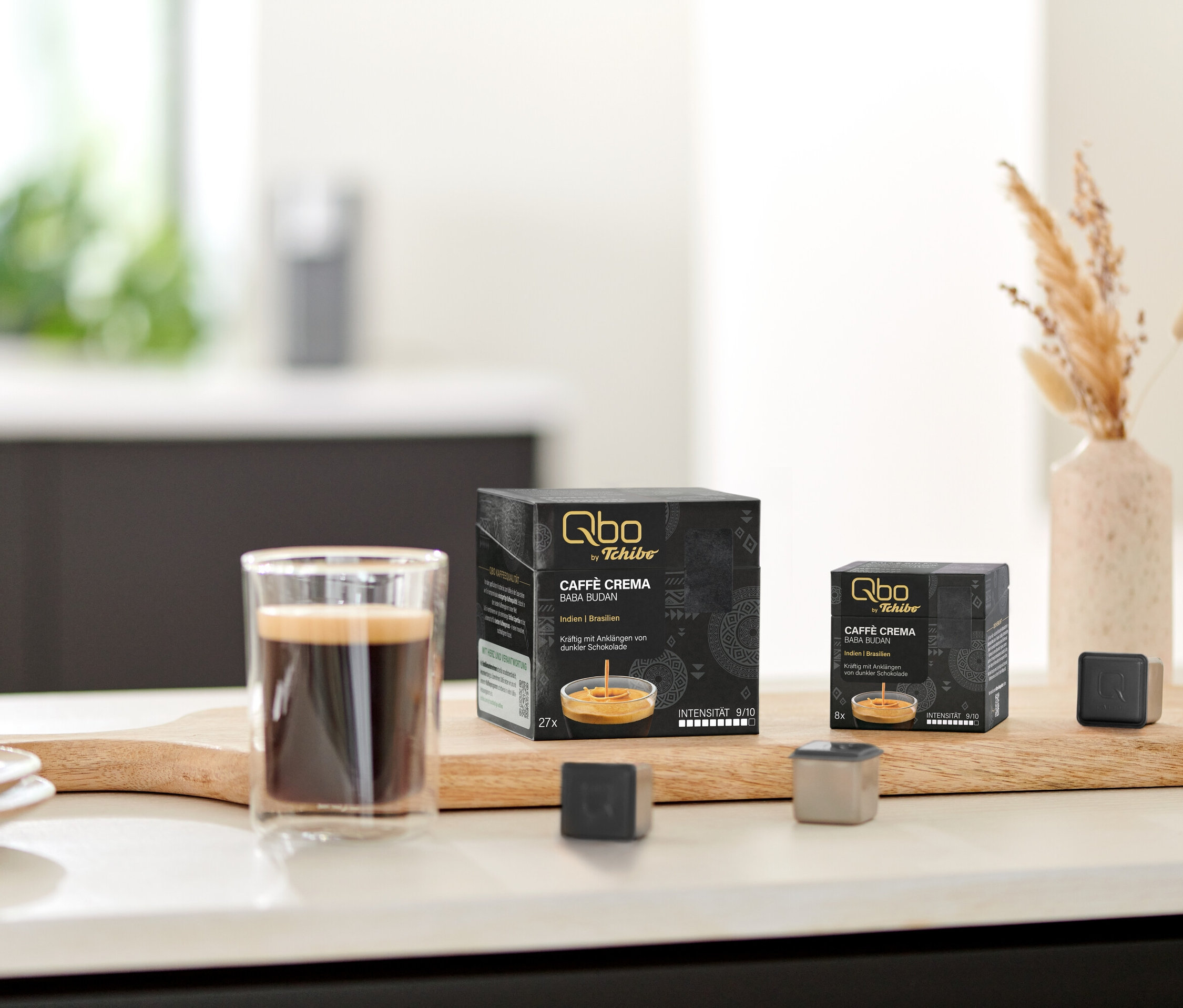 Auf einem Tisch stehen Qbo Caffè BABA BUDAN Kapseln, eine Tasse Kaffee und Kapsel-Prototypen auf einem Holzbrett.