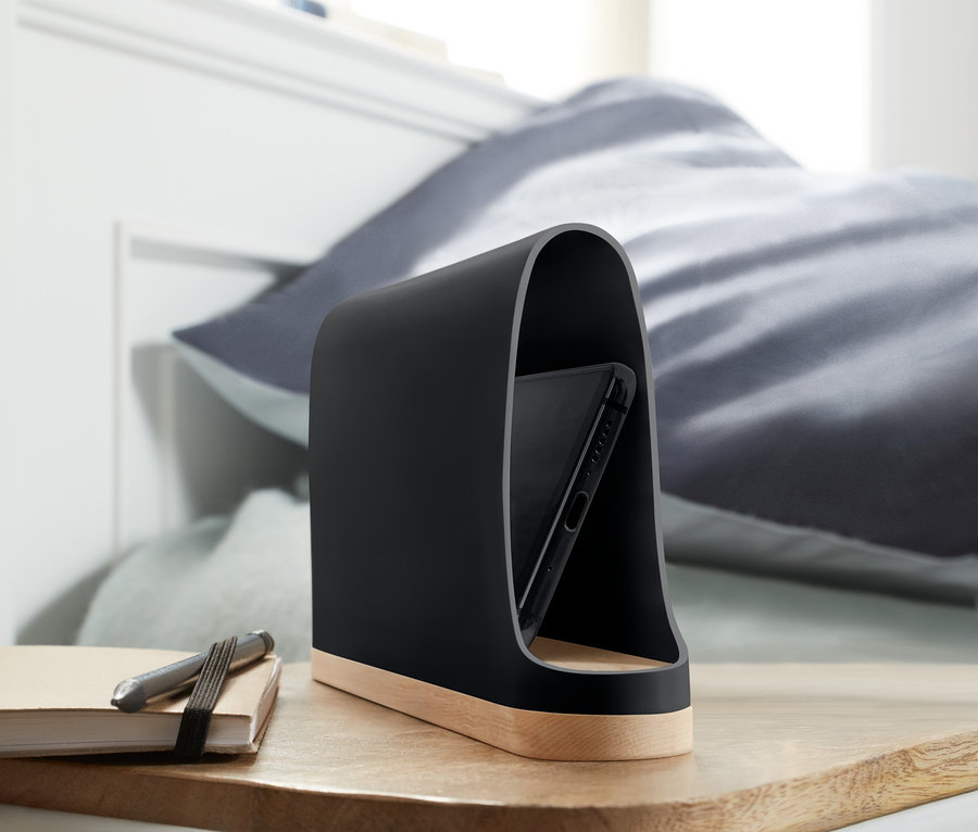 Handy in einer schwarzen strahlenabschirmenden Handyhalterung auf einem Holznachttisch neben einem Block und Stift.