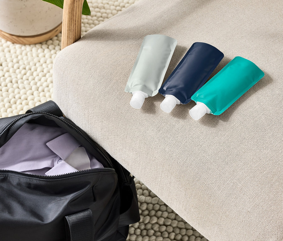 Drei wiederbefüllbare Reiseflaschen liegen auf einem beigen Sessel neben einer schwarzen Sporttasche und einer Topfpflanze.