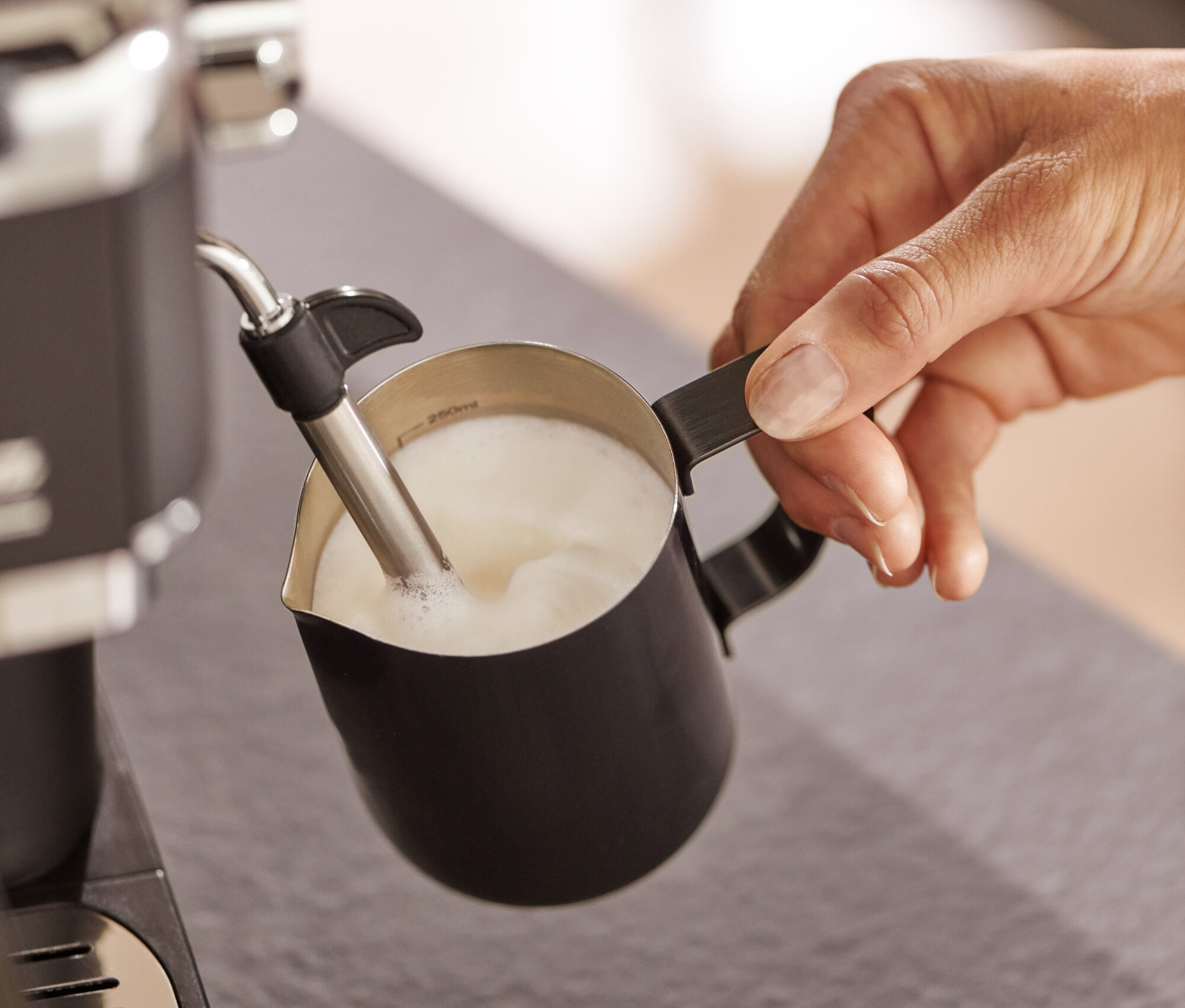 Person schäumt Milch in einem schwarzen Milchkännchen mit einer Kaffeemaschine auf.