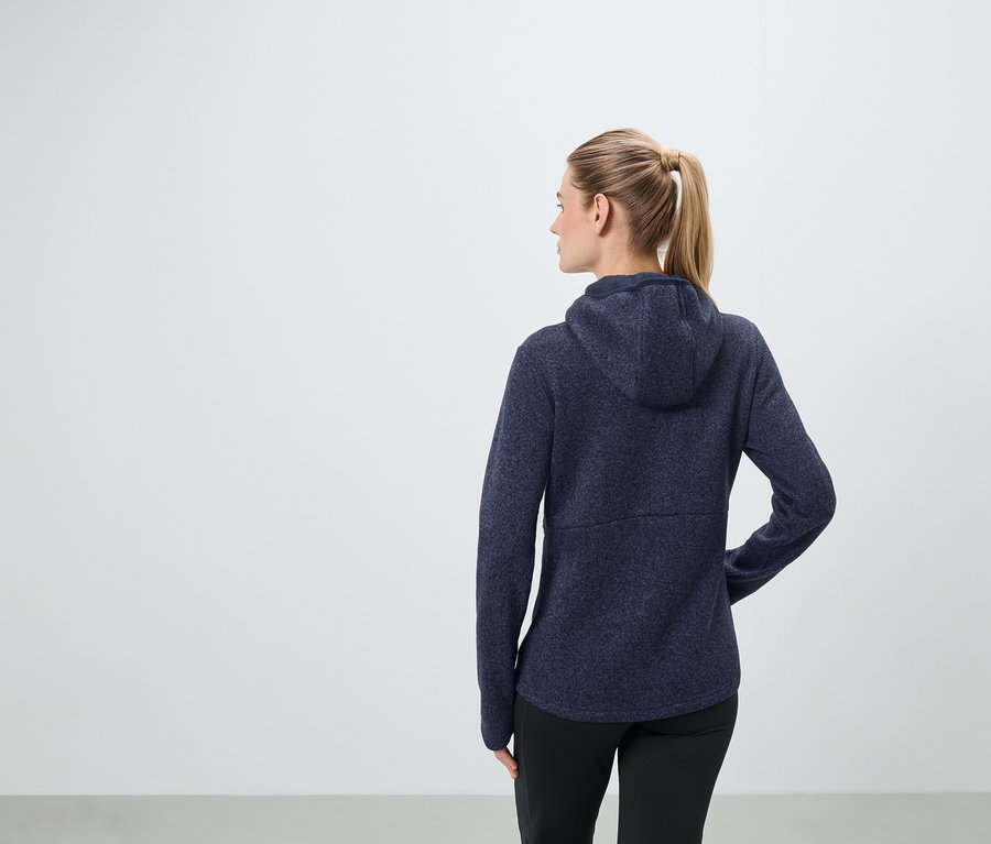 Frau mit blonden Haaren, die zu einem Pferdeschwanz zusammengebunden sind, trägt eine dunkelblaue Kapuzenjacke und schwarze Hosen.