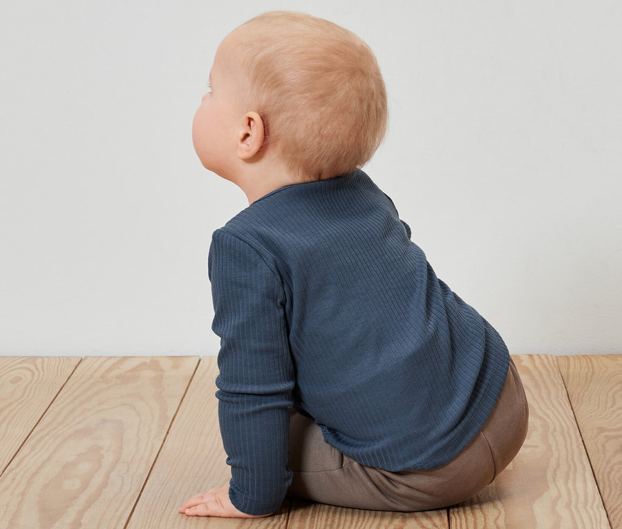 Ein Baby sitzt auf einem Holzboden und trägt ein blaues Langarmshirt mit Rippstruktur.