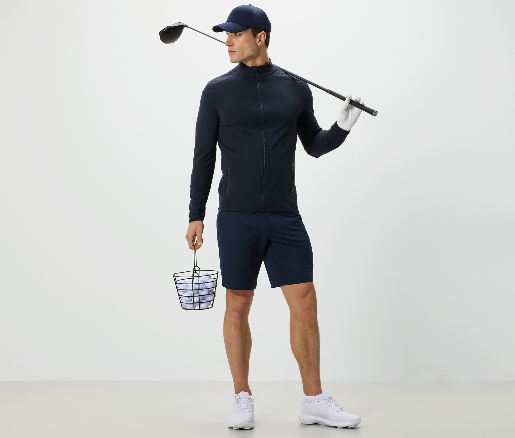 Mann in marineblauer Funktionsjacke hält Korb mit Golfbällen und Golfschläger.