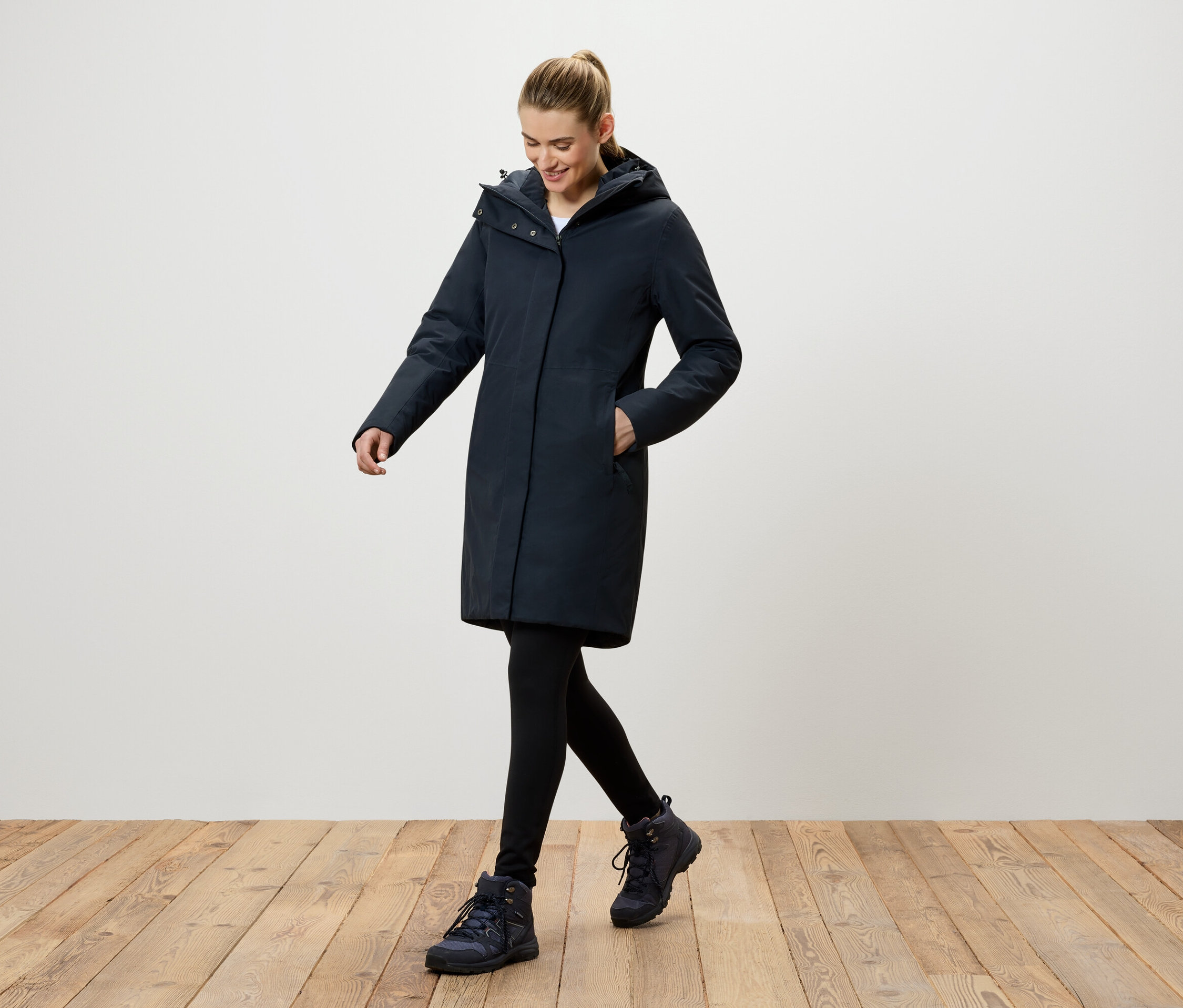 Eine Frau geht auf einem Holzboden und trägt einen dunkelblauen Mantel mit Kapuze, schwarze Leggings und dunkelblaue Schuhe.