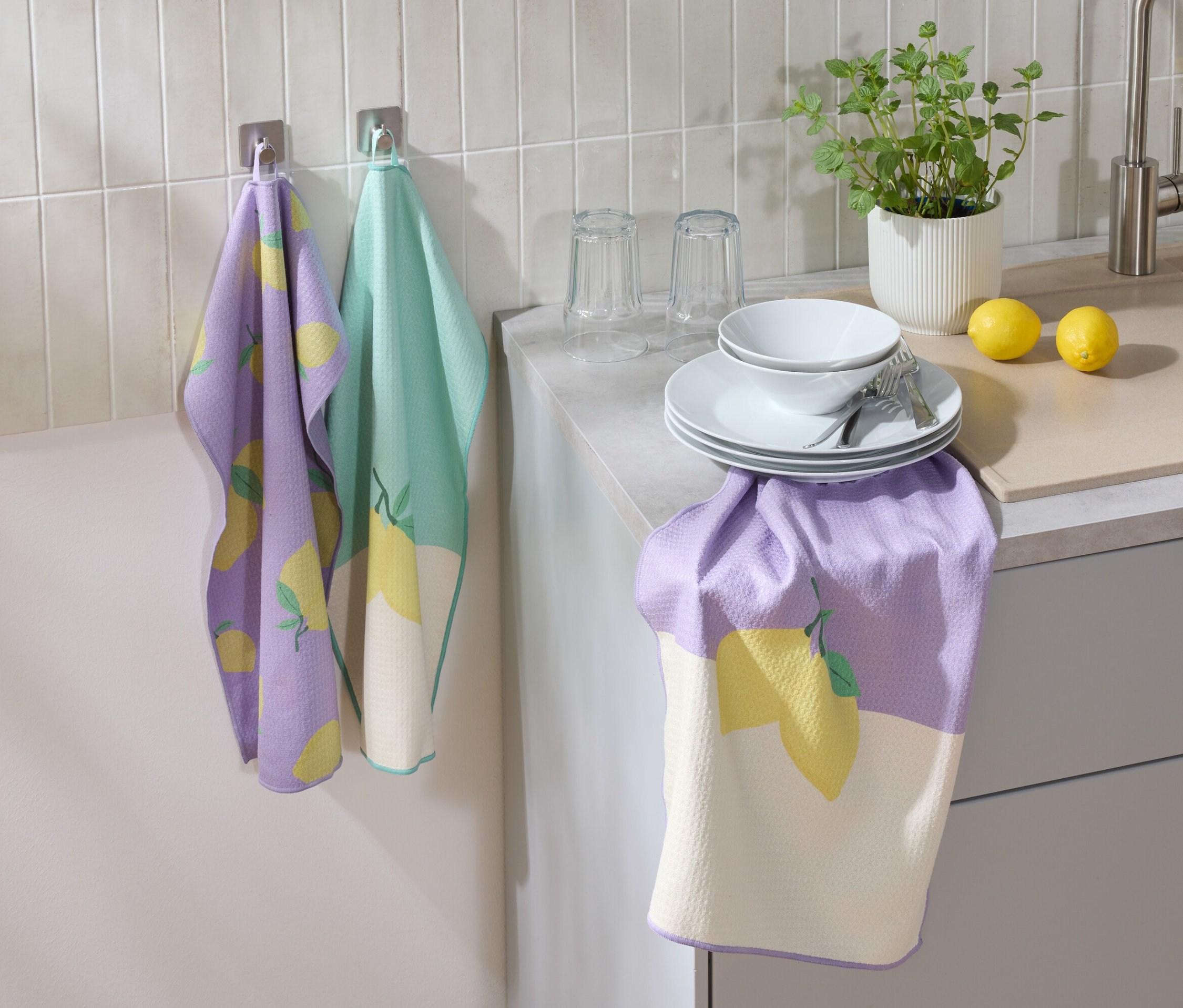 Ein lila und grünes Geschirrtuch mit Zitronenmuster hängt an der Wand. Auf der Arbeitsfläche stehen Teller, eine Schüssel, Gläser, Besteck, eine Zitrone und eine Blume.