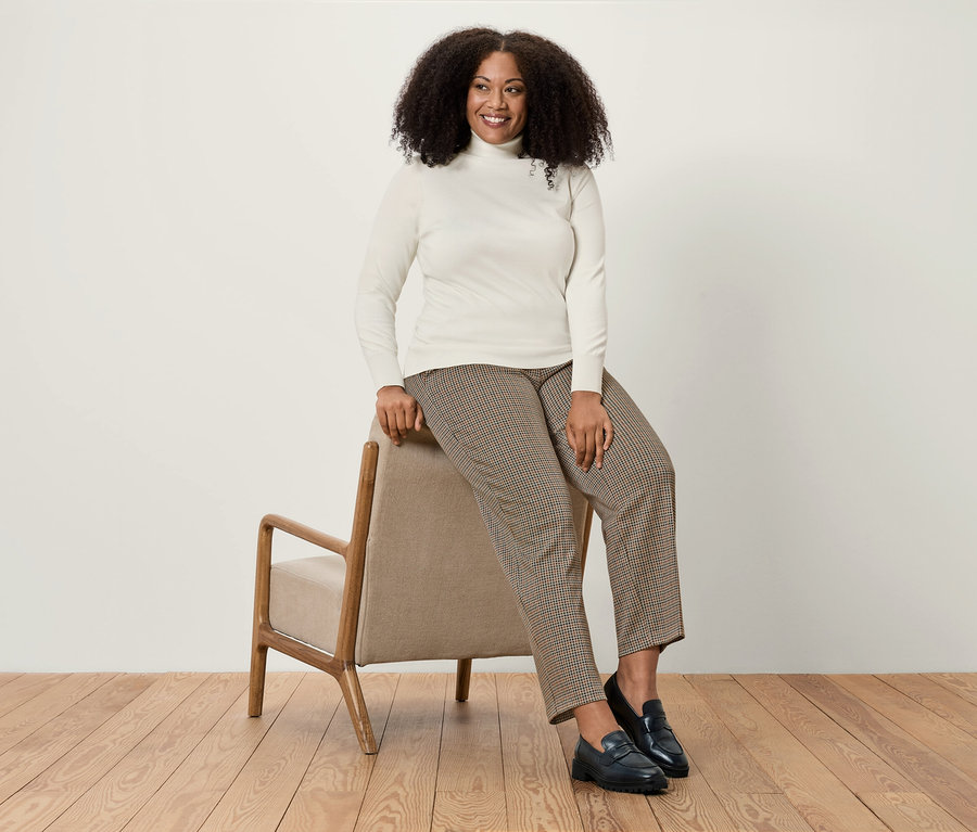 Eine Frau sitzt auf einem beigen Sessel und trägt einen weißen Rollkragenpullover, eine braune Hahnentritt-Hose und schwarze Slipper.