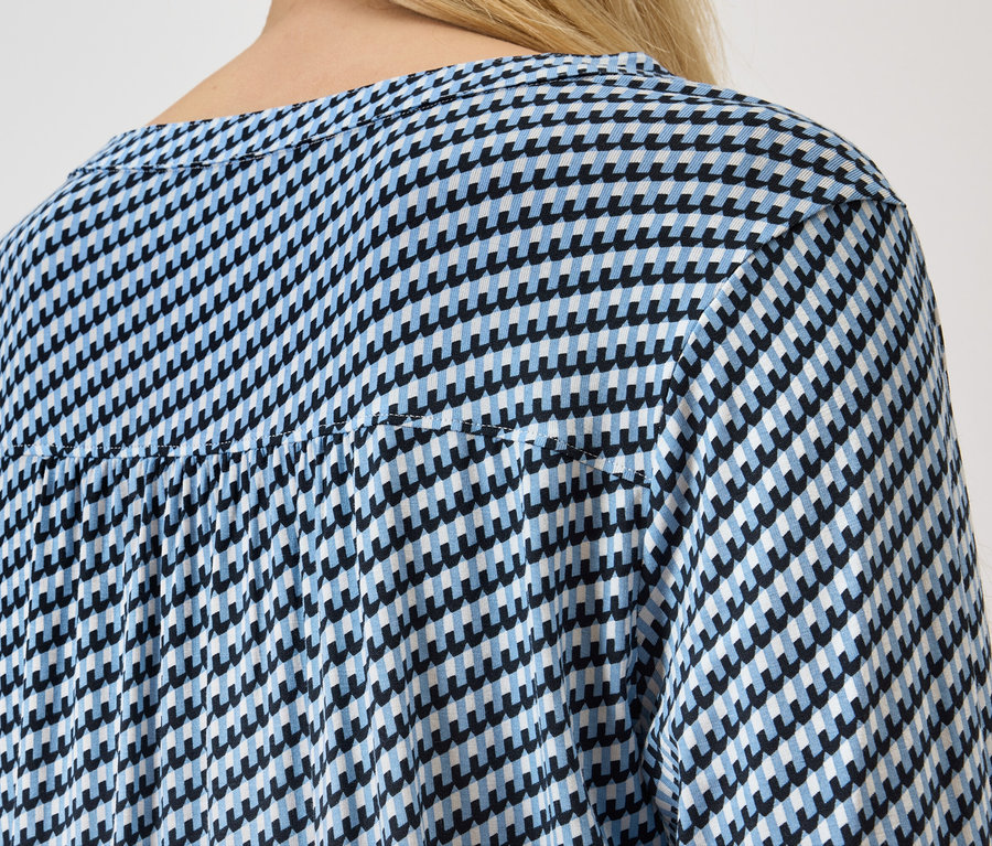 Rückansicht einer Frau mit blonden Haaren, die eine blau-weiße Hahnentrittbluse trägt.