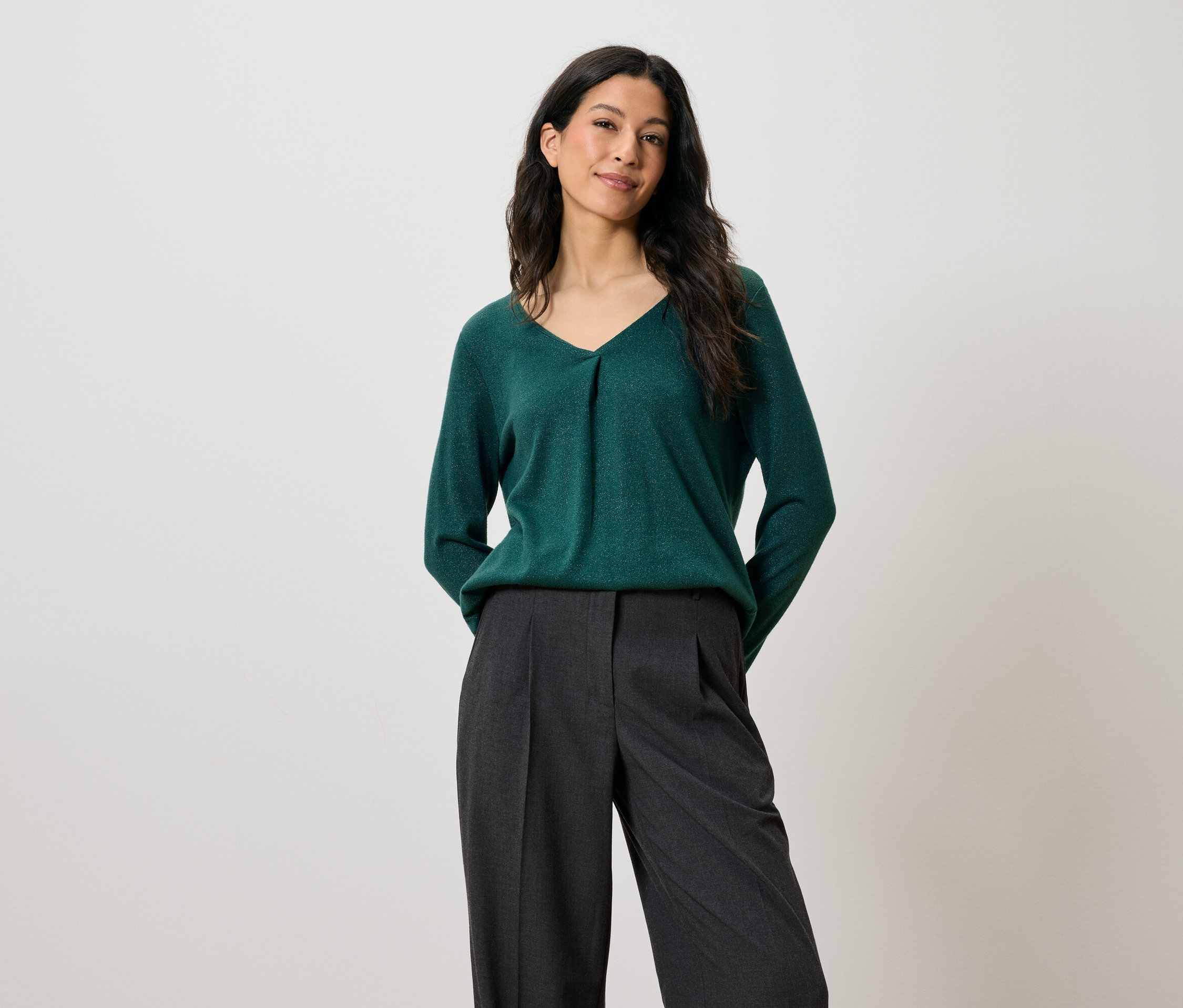 Frau mit dunklem Haar posiert in einem grünen Pullover und grauen Hosen.