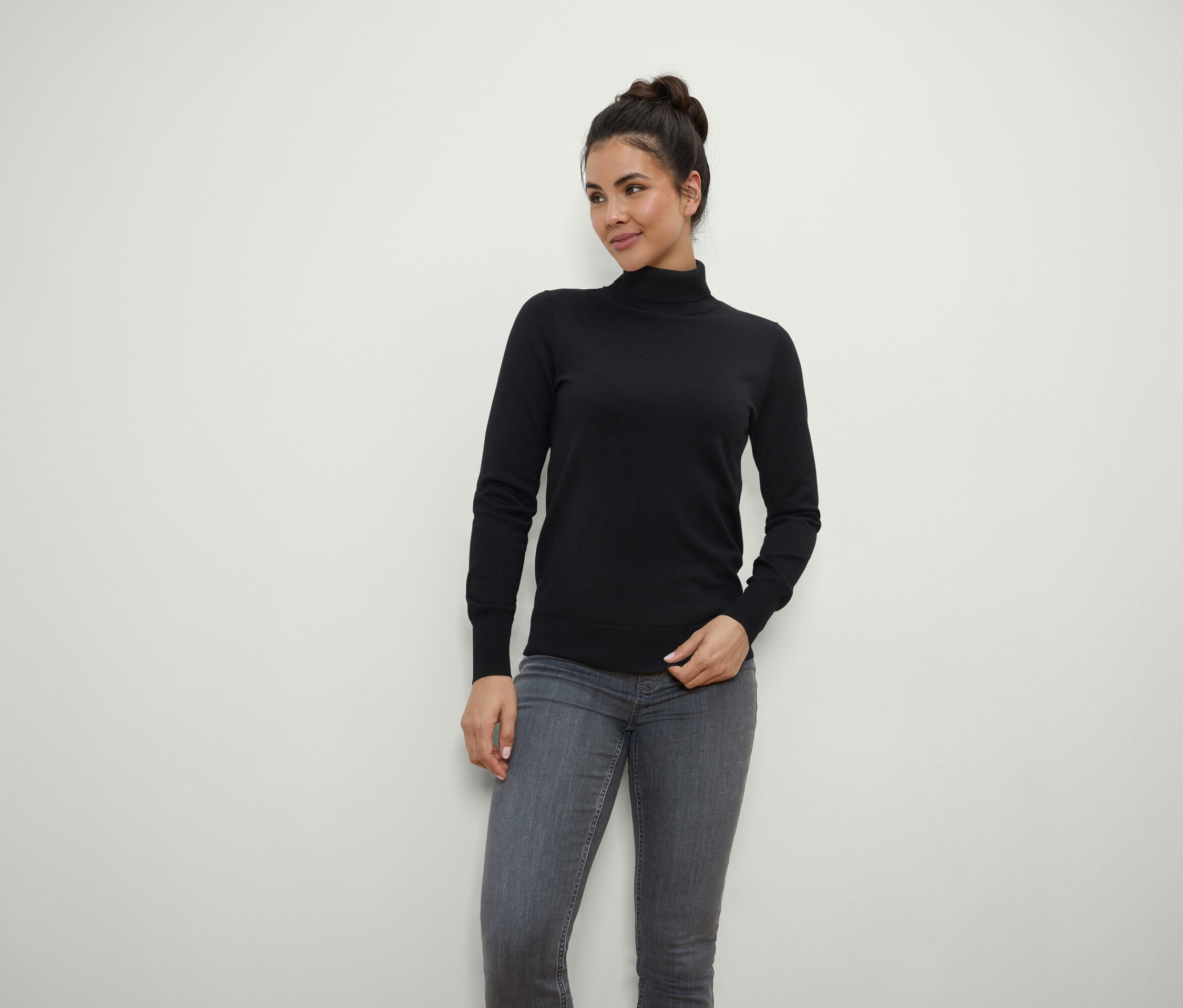 Frau mit braunen Haaren zum Dutt hochgesteckt posiert in schwarzem Rollkragenpullover und grauen Jeans.