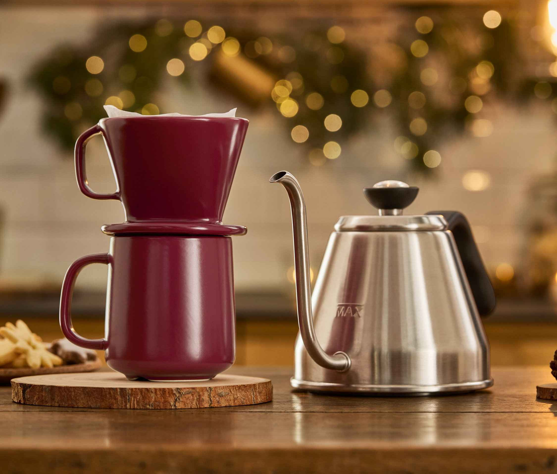 Ein Schwanenhalskessel steht neben einem beerenfarbenen Kaffeebecher mit Filter auf einem Holzsockel.