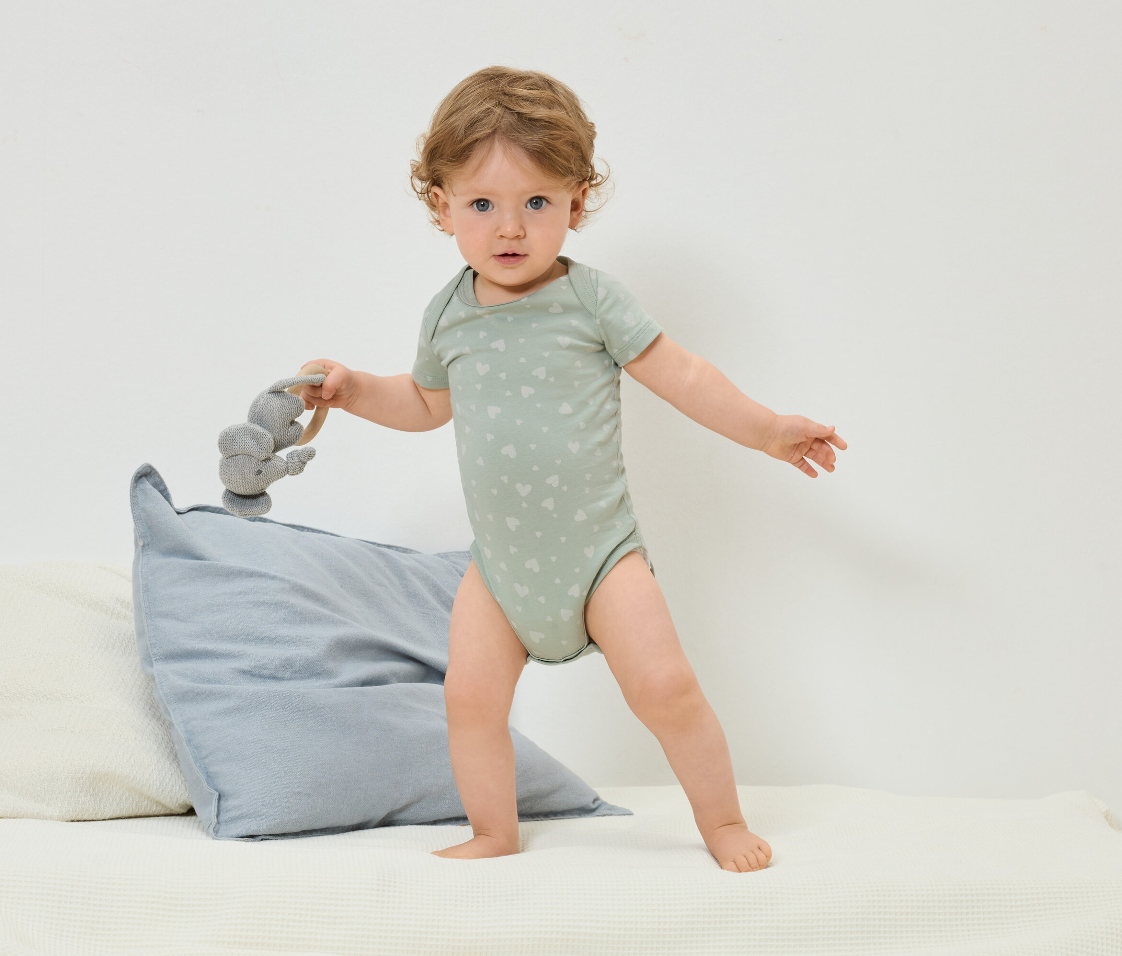 Ein Kleinkind in einem grünen Body mit Herzmuster steht auf einem Bett und hält ein graues Elefantenspielzeug.