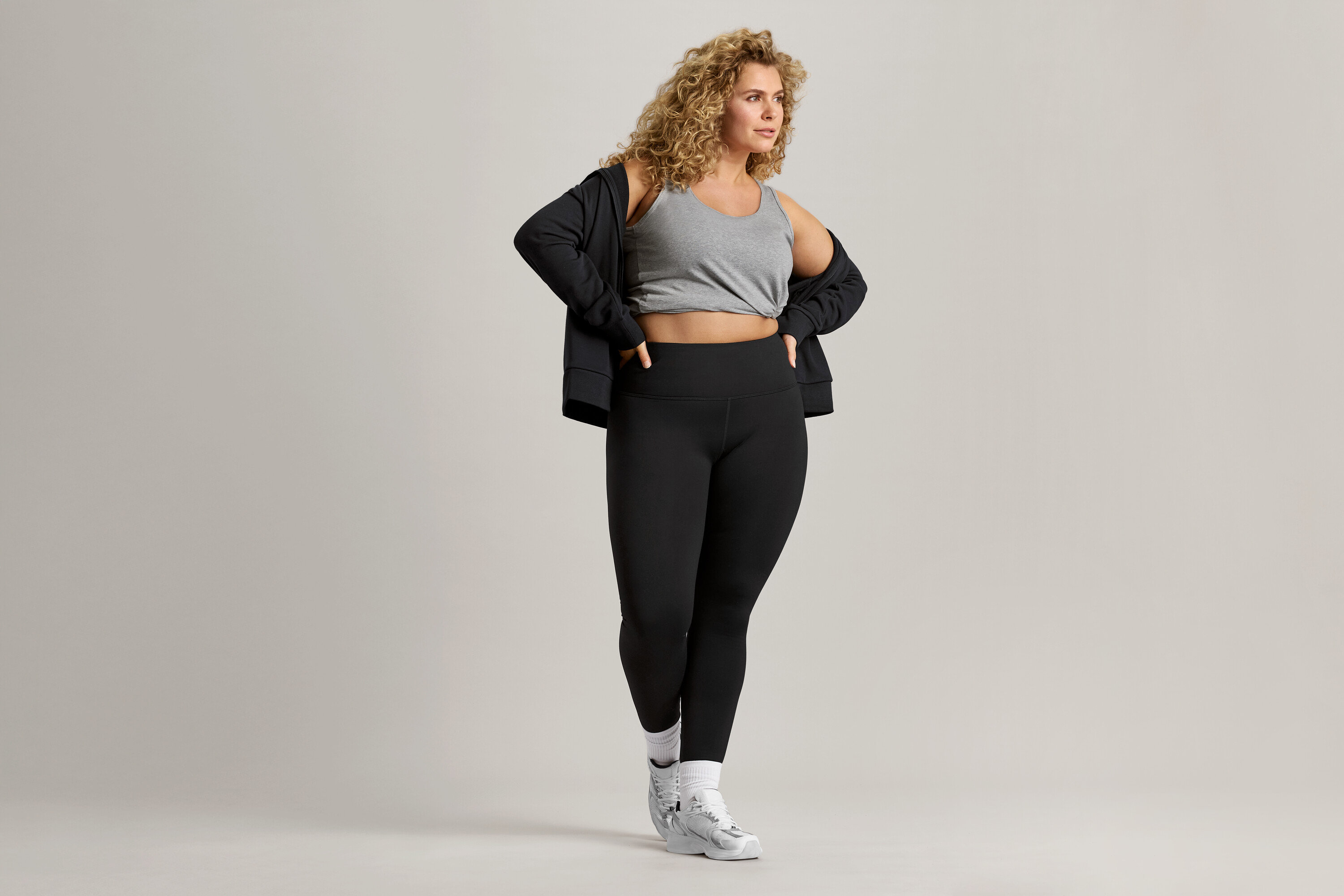 Frau mit lockigem Haar trägt ein graues Tanktop, eine schwarze Jacke und schwarze Leggings.