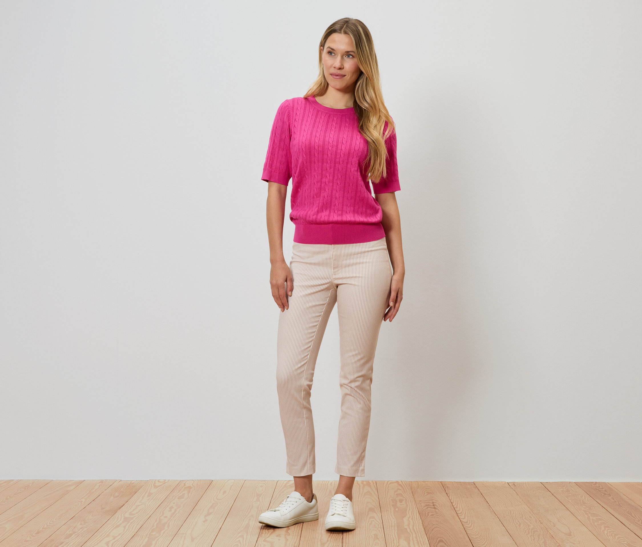 Frau steht vor einer Wand, trägt einen pinken Pullover, eine gestreifte Hose und weiße Schuhe.