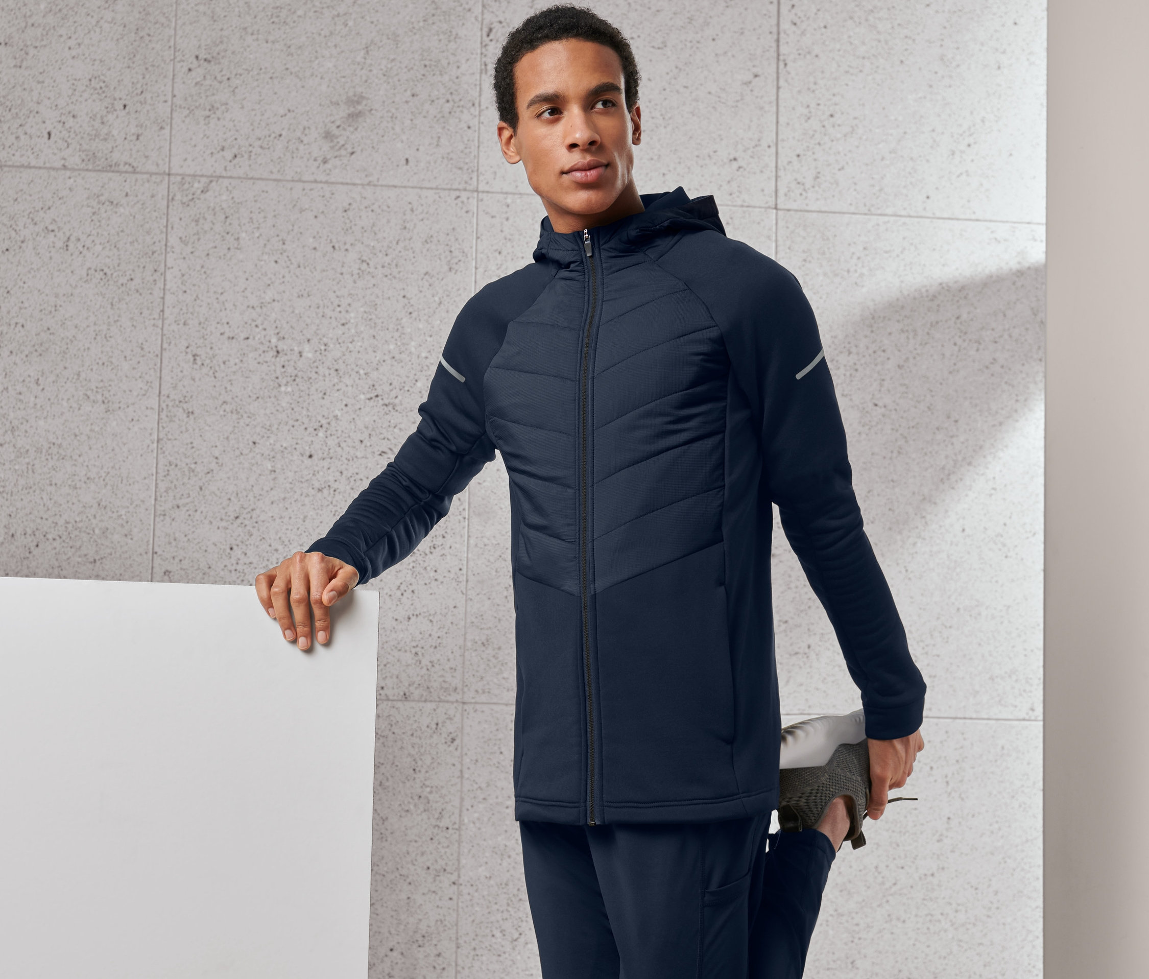 Mann dehnt sein Bein und trägt eine blaue Thermojacke.