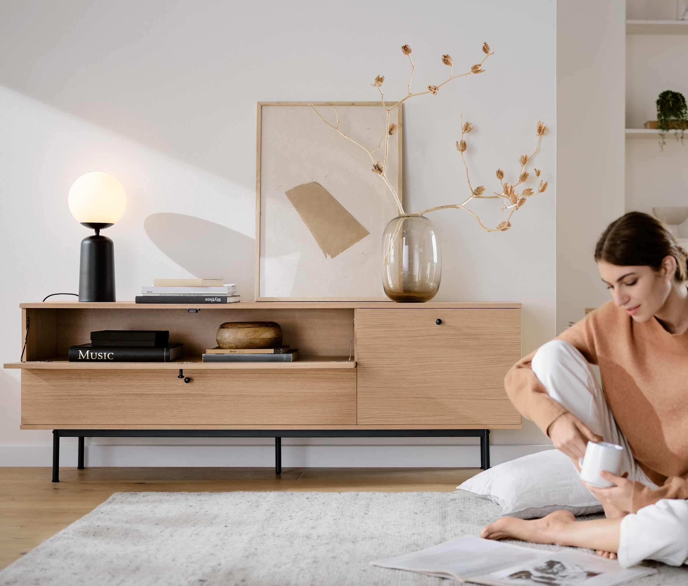 Eine Frau sitzt auf dem Boden neben einem Lowboard »Finnsbo« und einer Tischleuchte mit Glaskugel, schwarz.