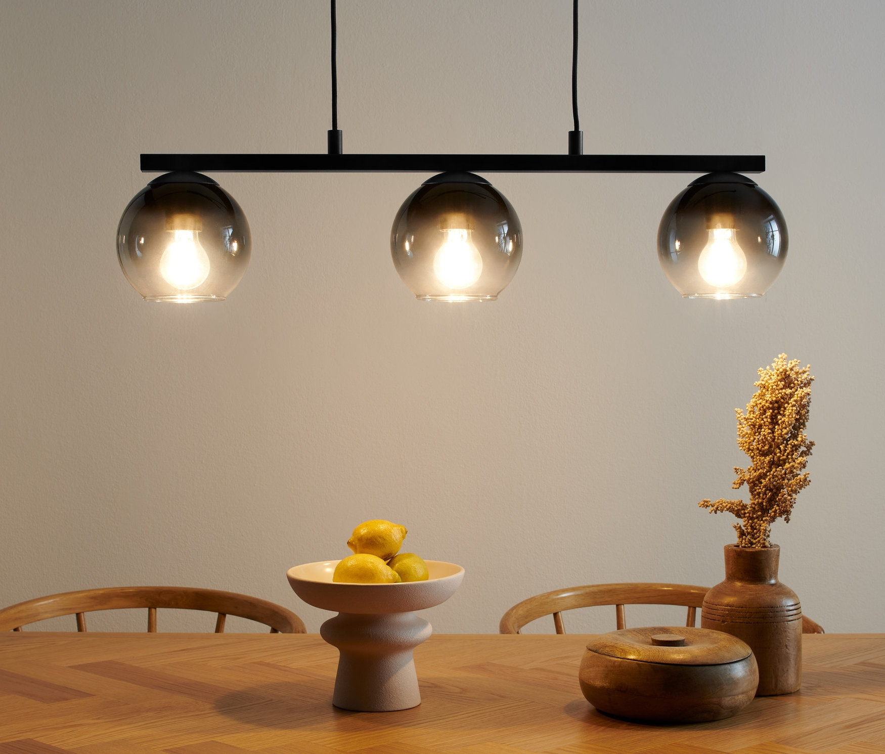 Pendelleuchte mit Rauchglas-Schirmen über einem Holztisch. Auf dem Tisch liegen eine Schale mit Früchten und eine Holzdose.