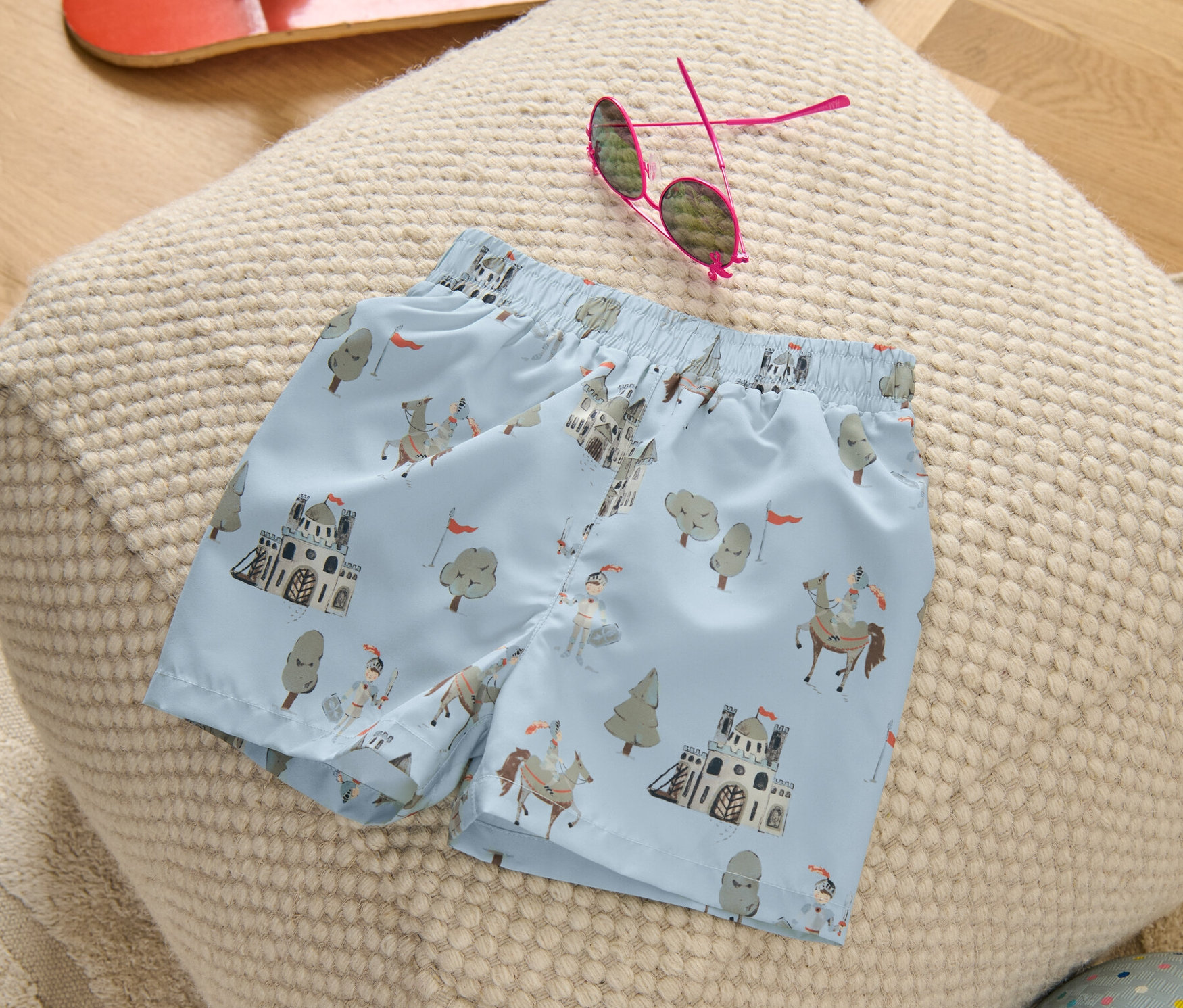 Kinder-Boardshorts mit Rittermuster liegen auf einem Hocker, daneben ein Skateboard, eine Sonnenbrille und ein Ball.