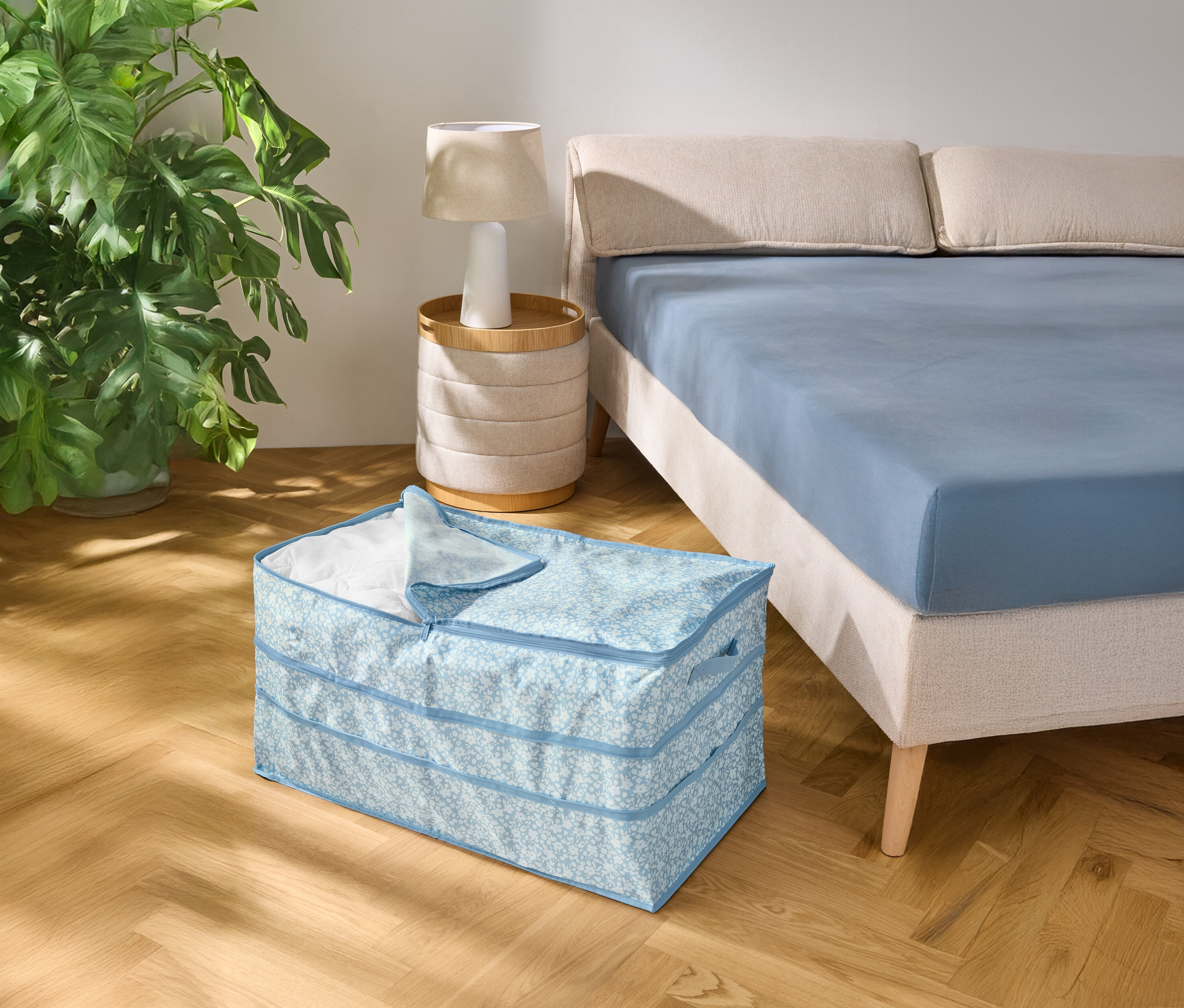 Auf einem Holzboden steht eine hellblau gemusterte Aufbewahrungsbox neben einem Bett mit blauem Bettlaken.