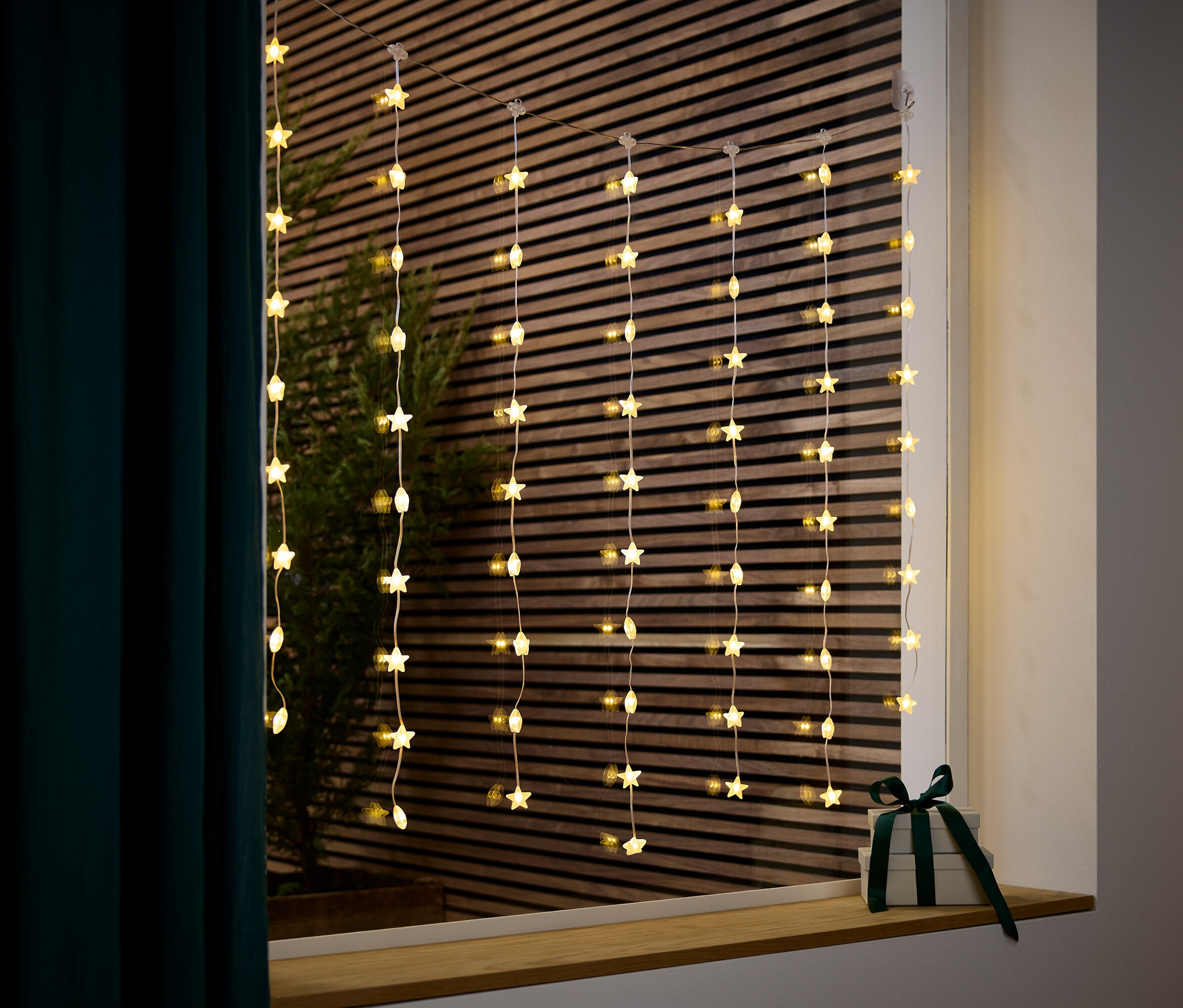 Eine Lichterkette mit Sternen hängt in einem Fenster neben einem Geschenk mit einem dunkelgrünen Band.