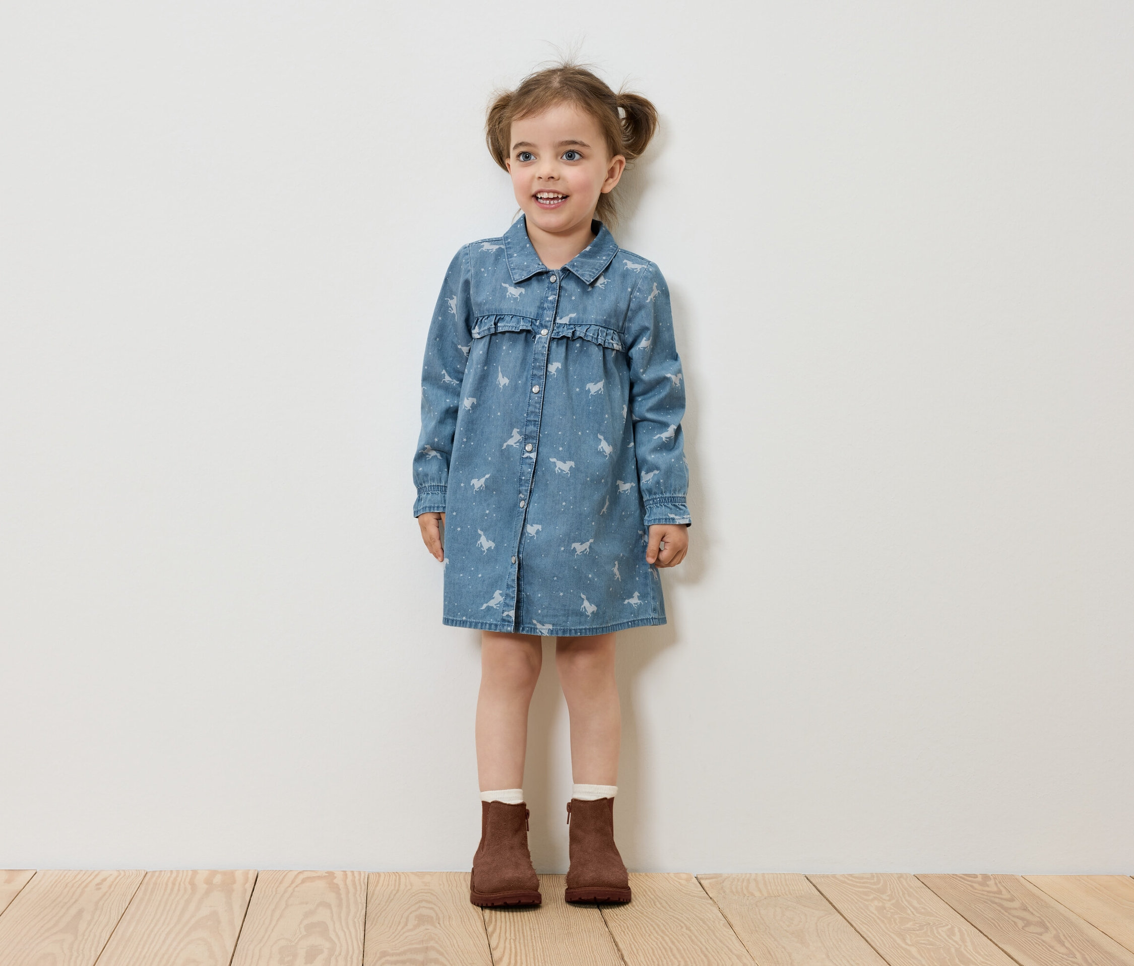 Ein kleines Mädchen steht auf einem Holzboden und trägt ein Jeanskleid mit Pferdeaufdruck und braune Stiefel.