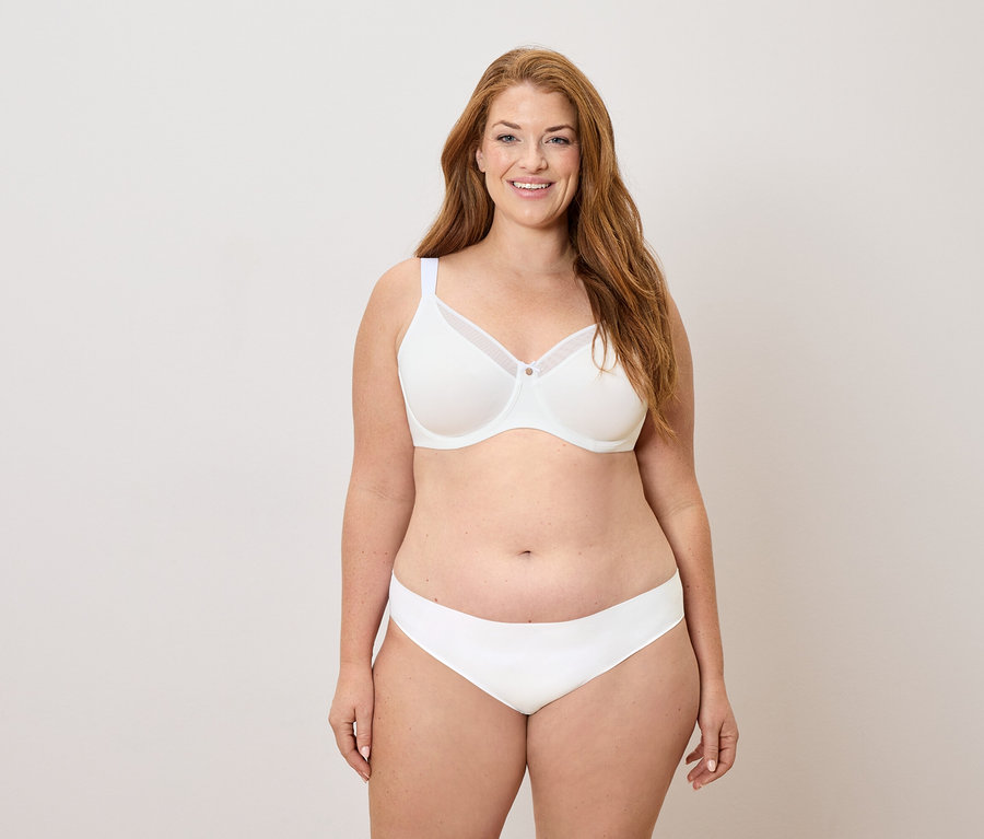 Frau mit langen roten Haaren, die einen weißen BH und Slip trägt und lächelt.