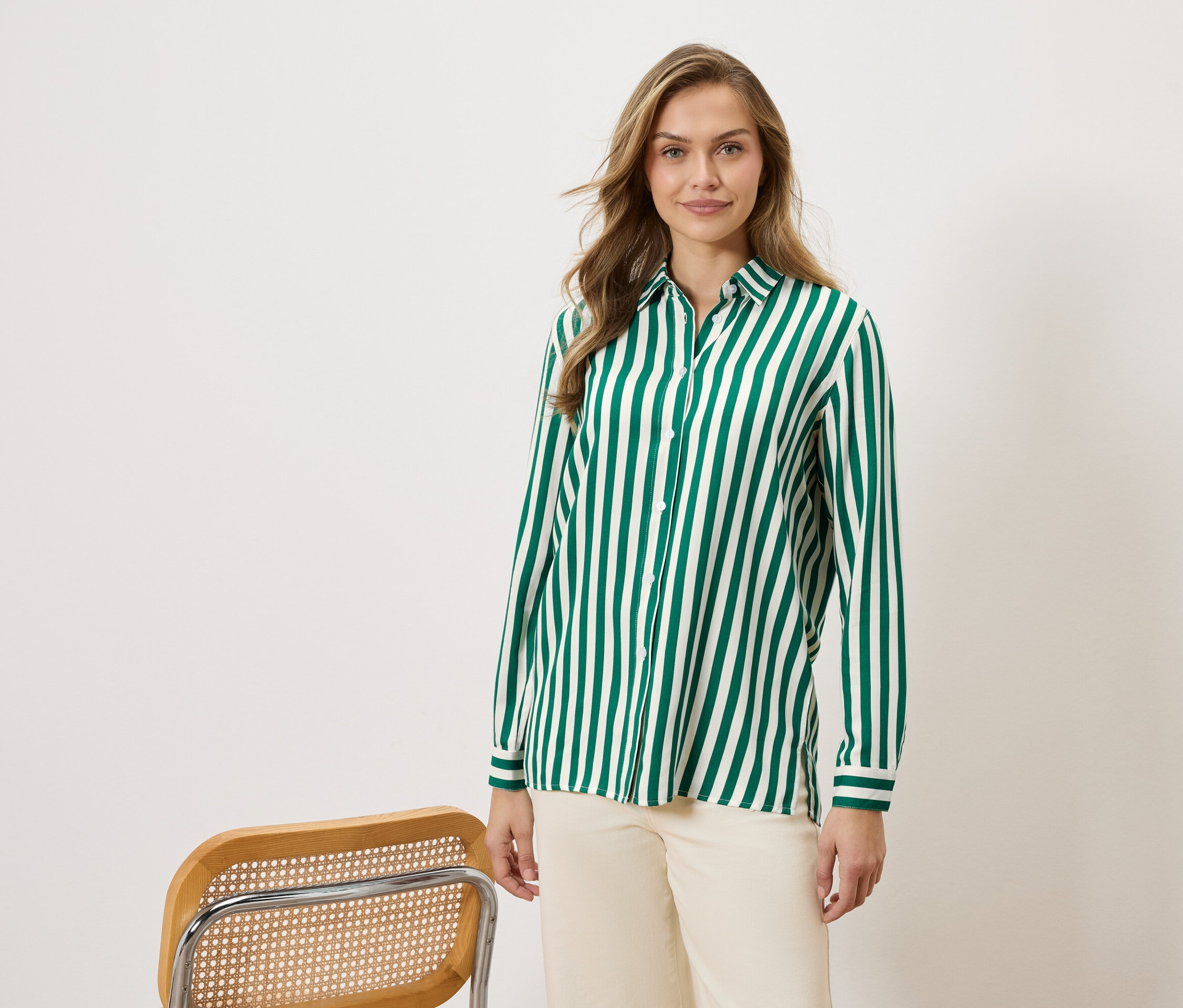 Frau trägt ein grün-weiß gestreiftes Hemd und beigefarbene Hosen, neben einem Stuhl.