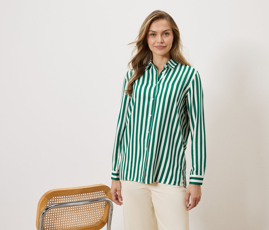 Frau trägt ein grün-weiß gestreiftes Hemd und beigefarbene Hosen, neben einem Stuhl.