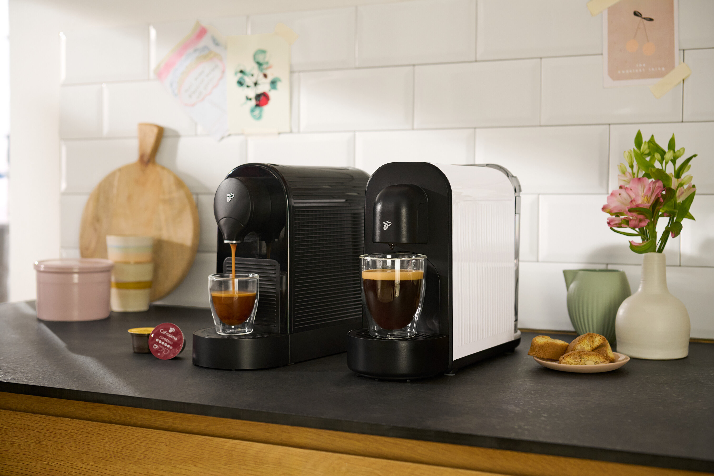 Auf einer Küchentheke stehen zwei Cafissimo pure+, black Kaffeemaschinen, eine schwarze und eine weiße, wobei die schwarze Maschine gerade einen Espresso in eine Tasse brüht.