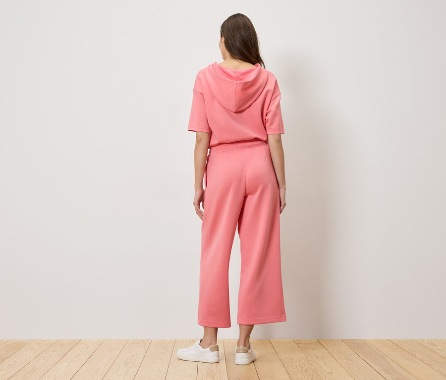 Eine Frau steht mit dem Rücken zur Kamera und trägt einen rosafarbenen Hoodie und eine Culotte Hose.