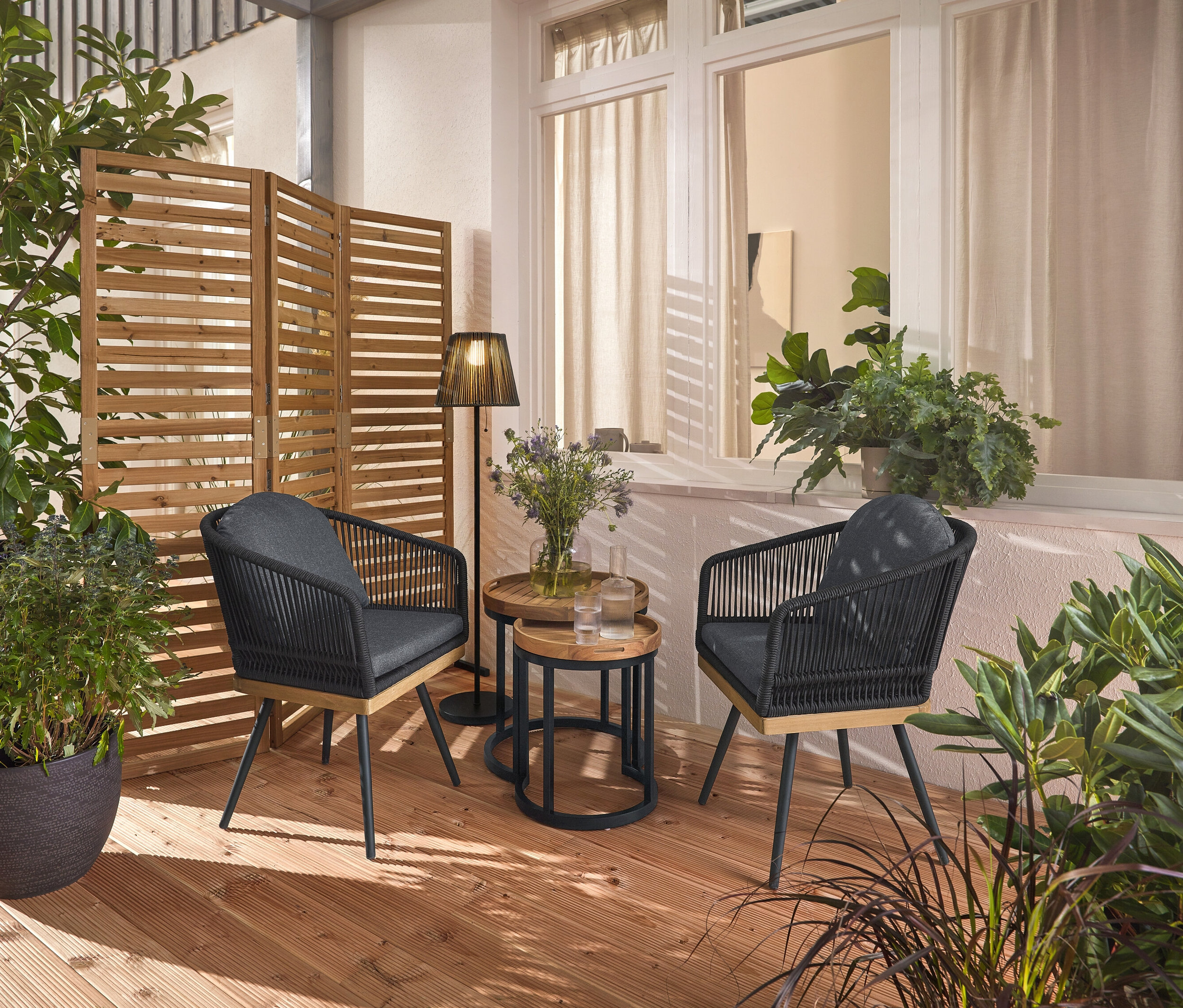 Zwei drehbare Gartenstühle mit Textilgeflecht stehen auf einer Holzterrasse, dazwischen zwei Garten-Beistelltische mit abnehmbaren Tabletts, Ø ca. 40 cm und 50 cm. Im Hintergrund ein flexibler Paravent und eine Akku-Solar-Stehleuchte.