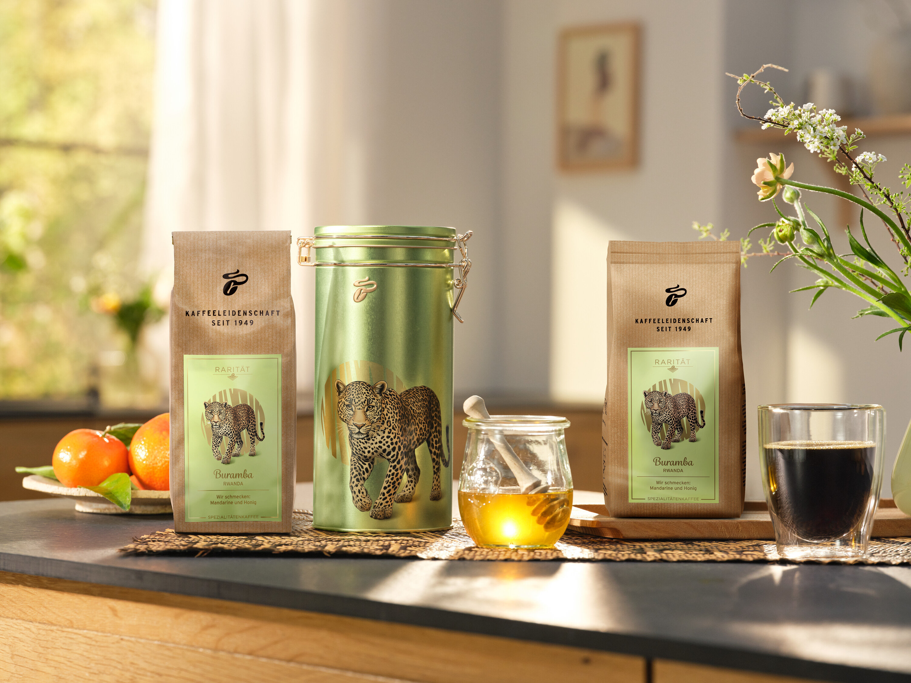 Auf einer Küchentheke stehen eine Kaffeedose und zwei Packungen »Buramba Rwanda« inkl. Kaffeedose neben einer Tasse Kaffee und Honig.