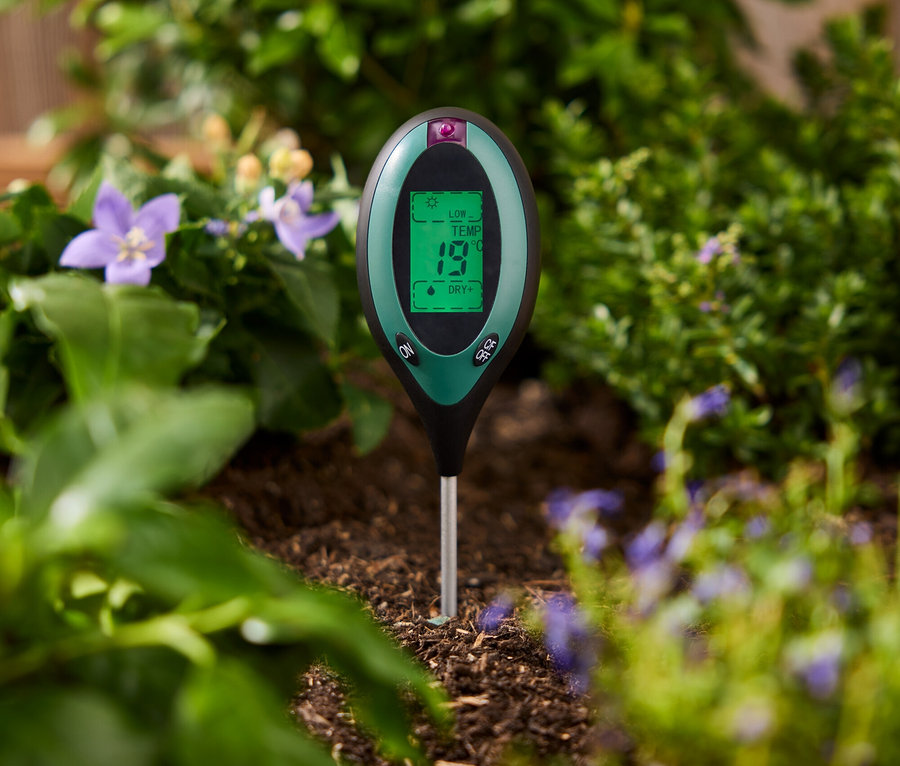 Ein grünes Bodentestgerät steckt im Boden zwischen Pflanzen.