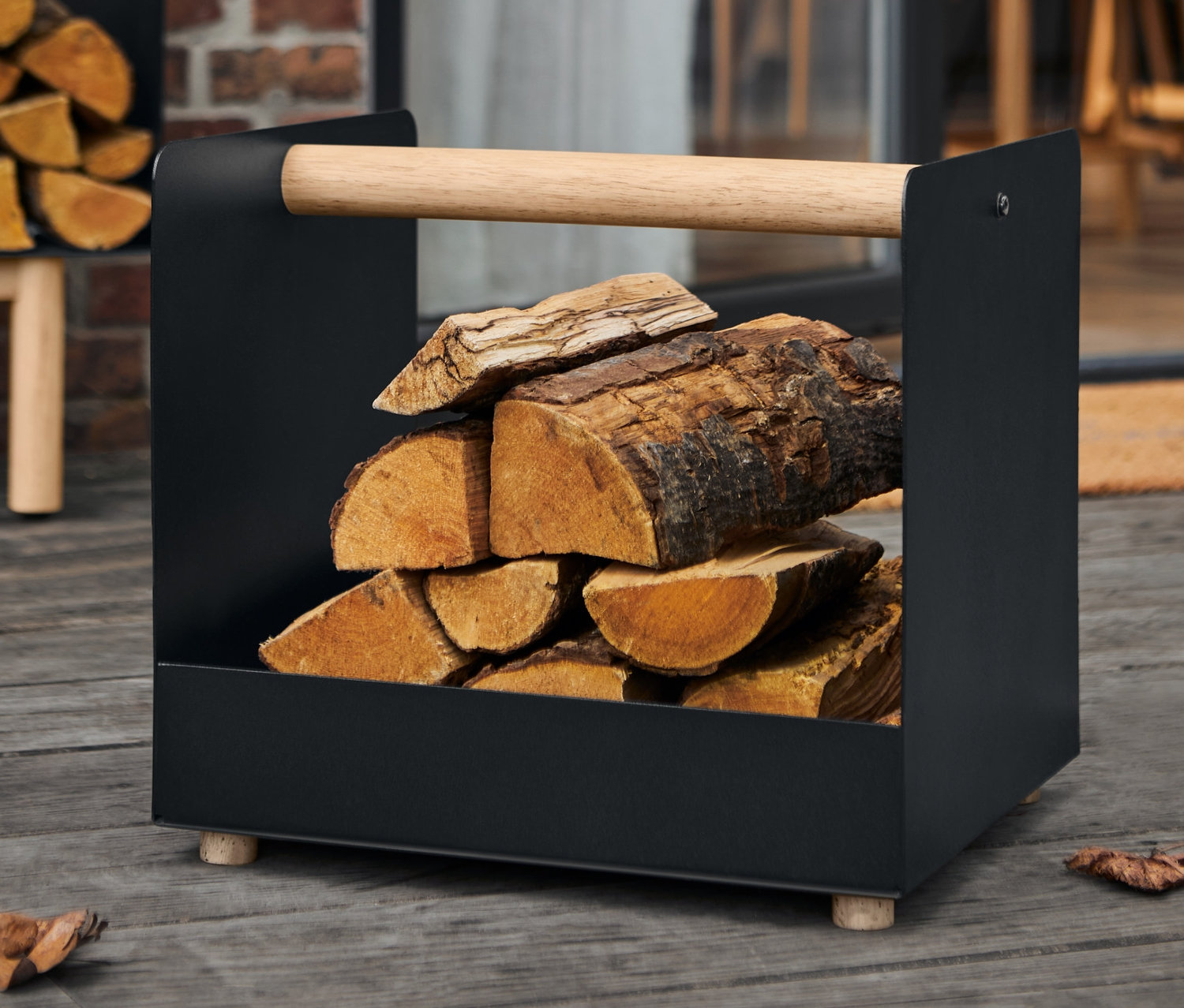 Ein schwarzer Kaminholz-Korb mit Holzgriff, gefüllt mit Holzscheiten. Im Hintergrund ist ein weiterer Kaminholzkorb und eine Ziegelwand zu sehen.