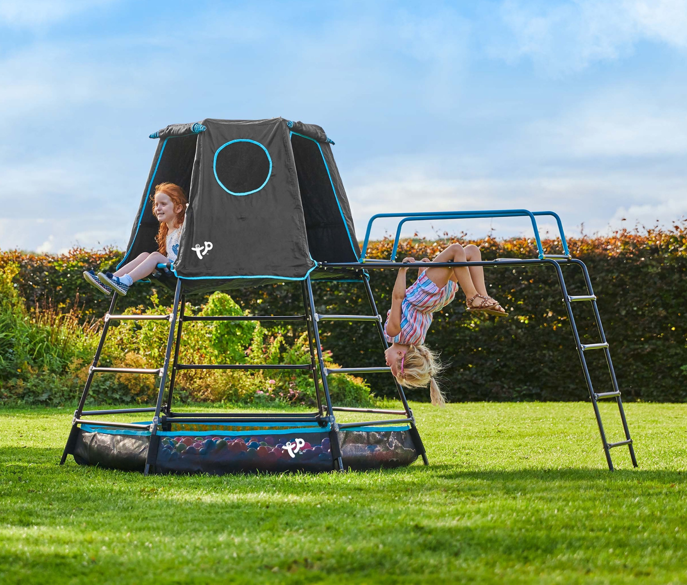Zwei Mädchen spielen auf dem Kletterturm »Explorer« mit Zelt auf einer Wiese.