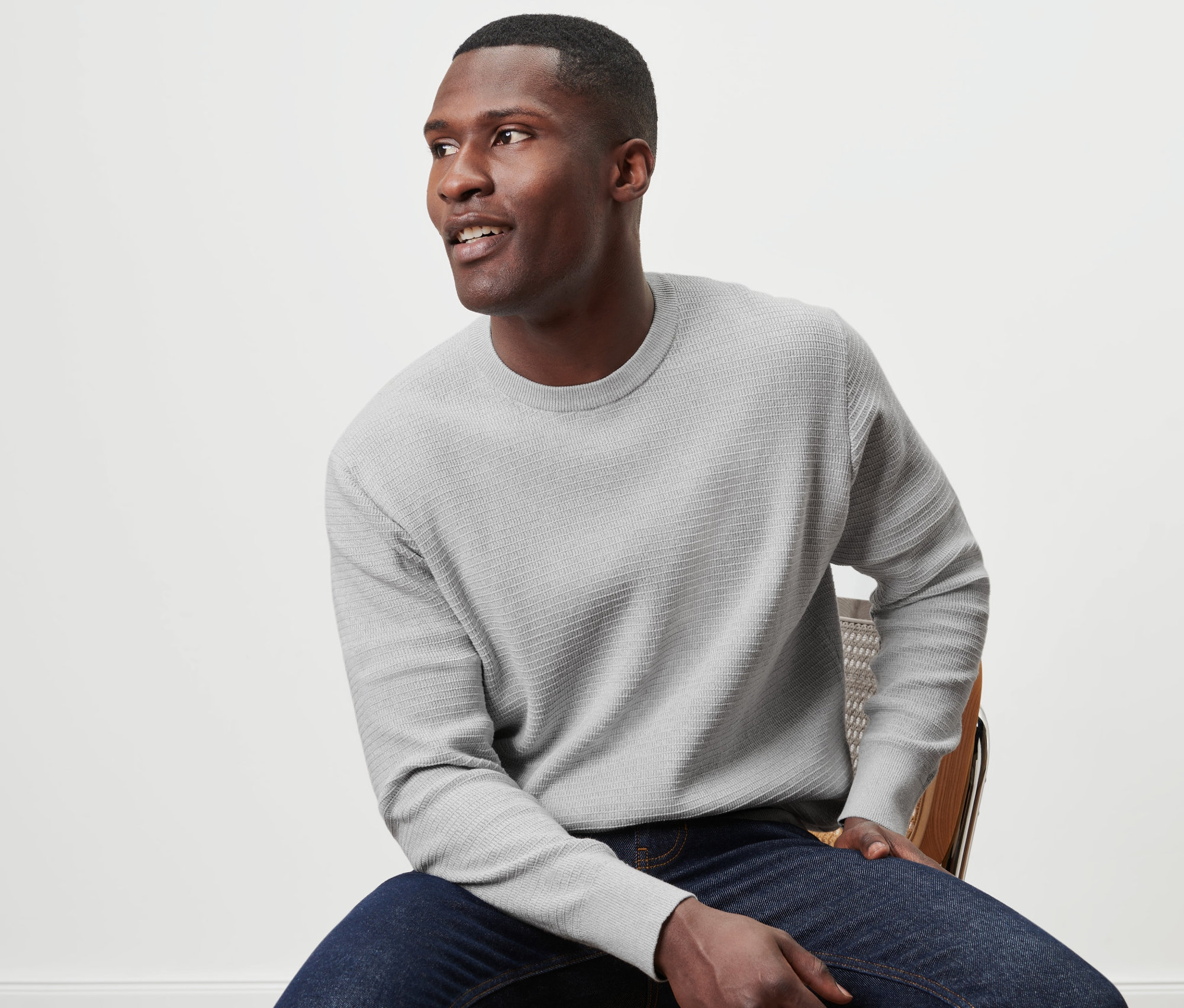 Mann sitzt auf einem Stuhl und trägt einen grauen Pullover mit Rundhalsausschnitt.