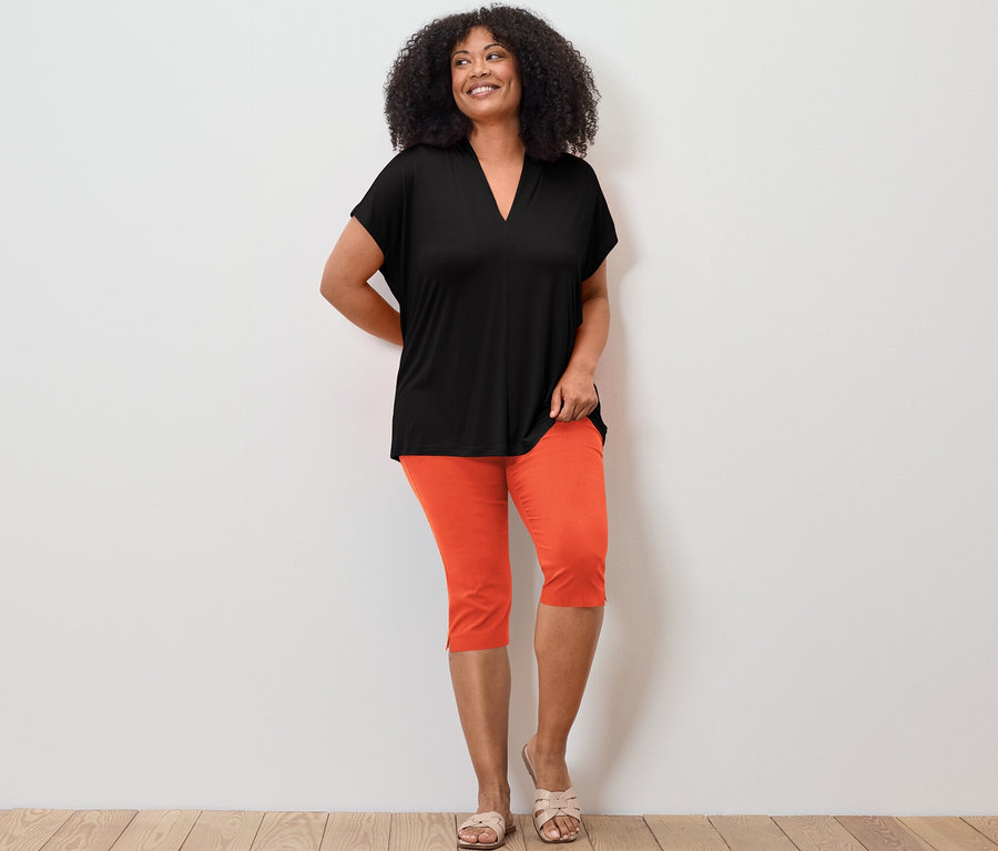 Frau posiert in Blusenshirt mit V-Ausschnitt, schwarz und 3/4-Stretchhose, orange.