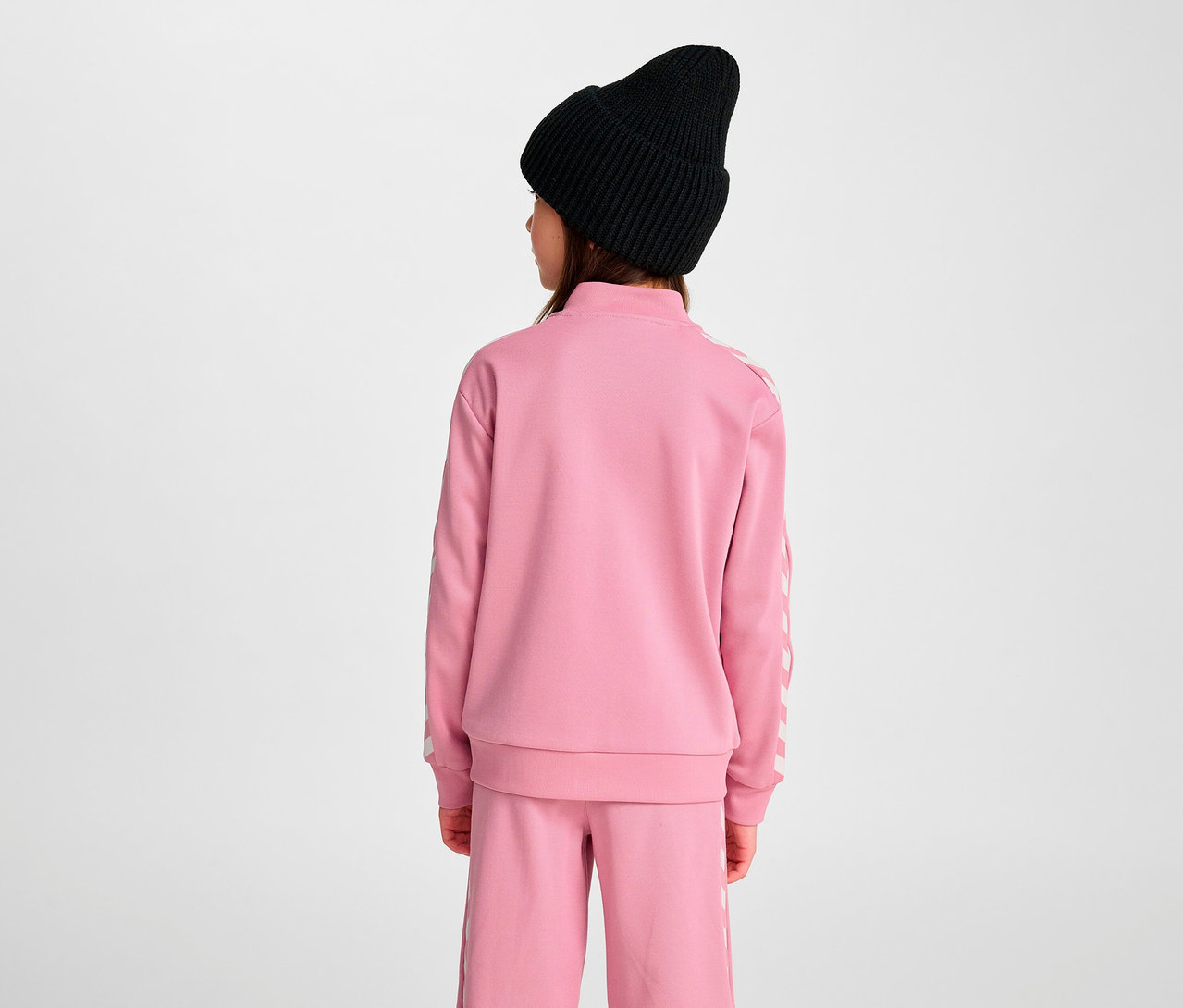 Mädchen von hinten trägt rosa Trainingsanzug und schwarze Mütze. Sie trägt eine HUMMEL HMLJR Archive Reg Poly Zip Jacket, rosa.