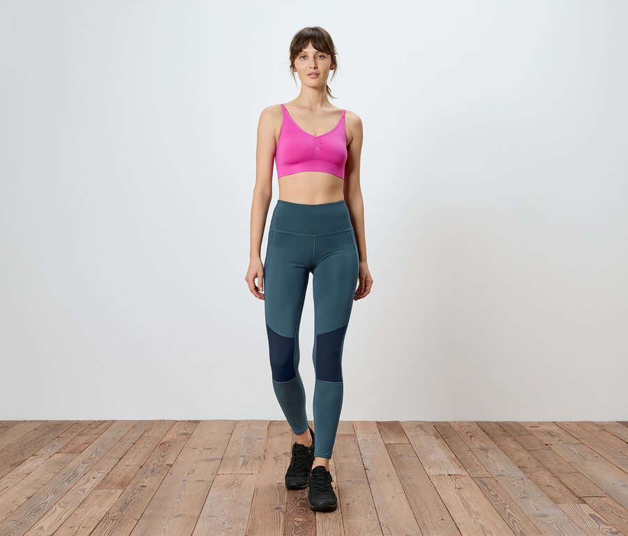 Frau geht auf Holzboden, trägt einen pinken Sport-BH und blaue Leggings mit dunklen Einsätzen.